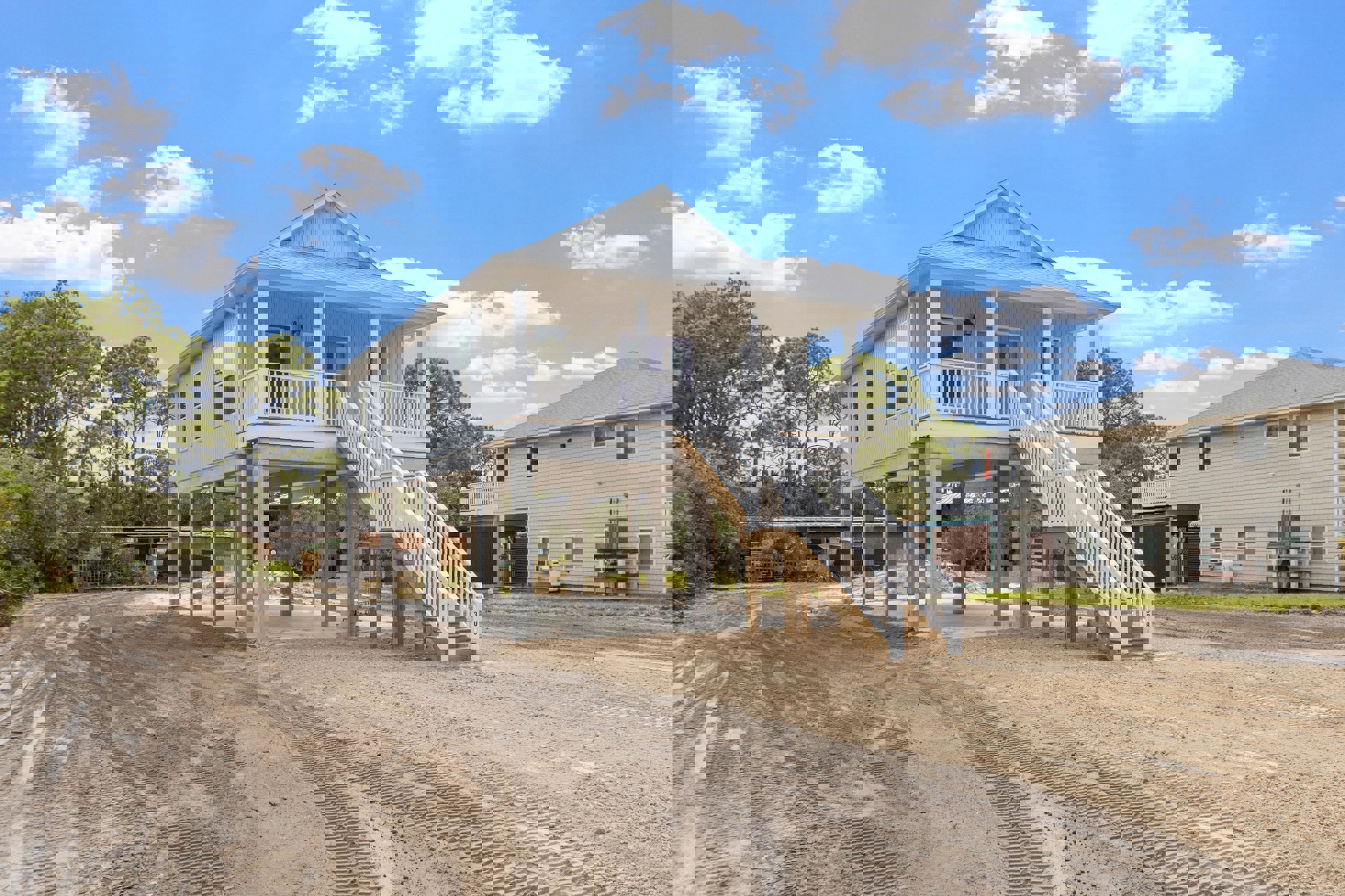 Two-story house with white siding, blue front door, covered porch, wooden stairs, attached garage, tire tracks on sandy driveway, windows, and blue sky with scattered clouds