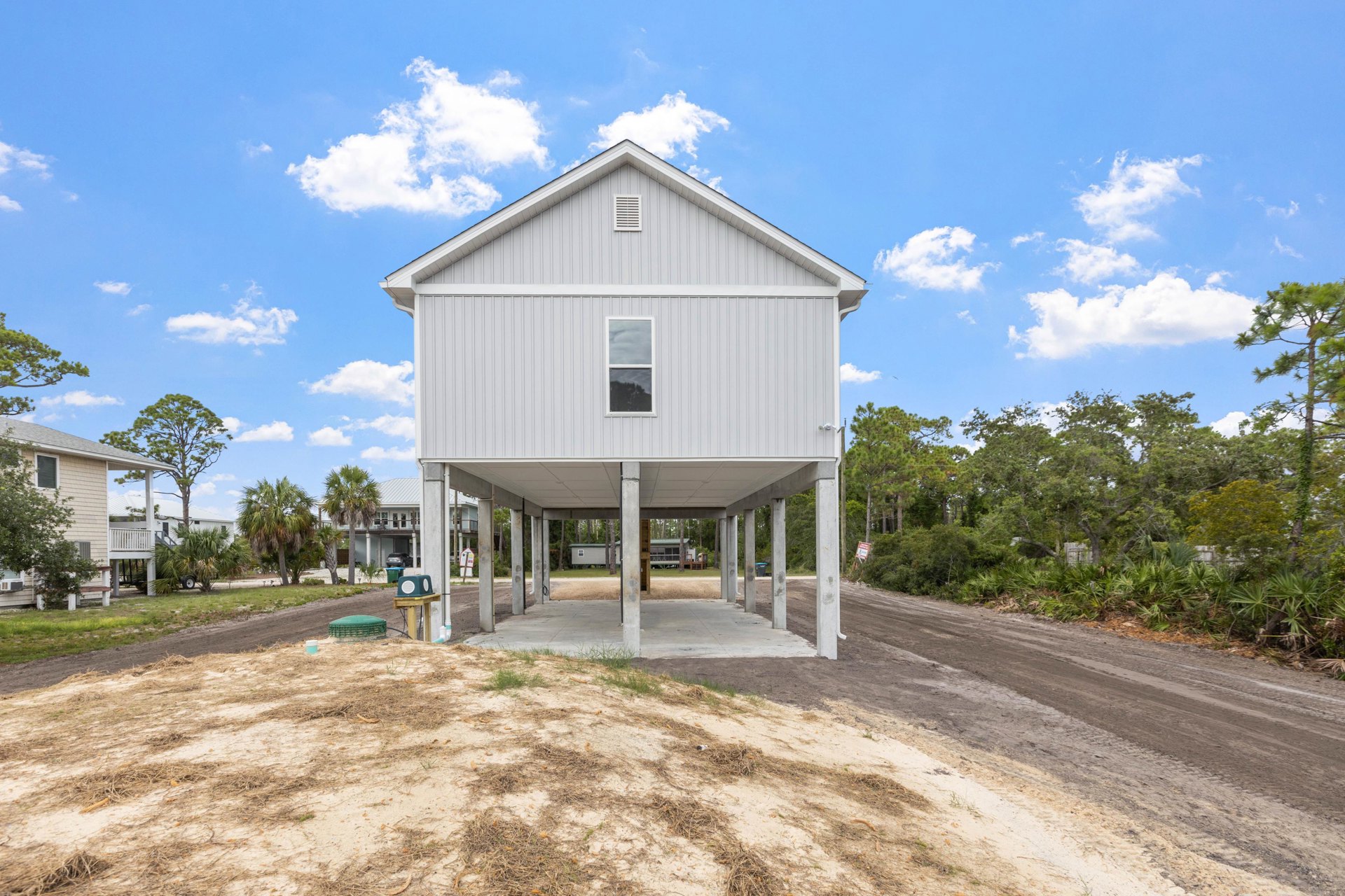 Elevated house on stilts with white-framed windows, attached garage, green utility box in front, situated on a dirt road under a cloudy grey sky, surrounded by sparse vegetation
