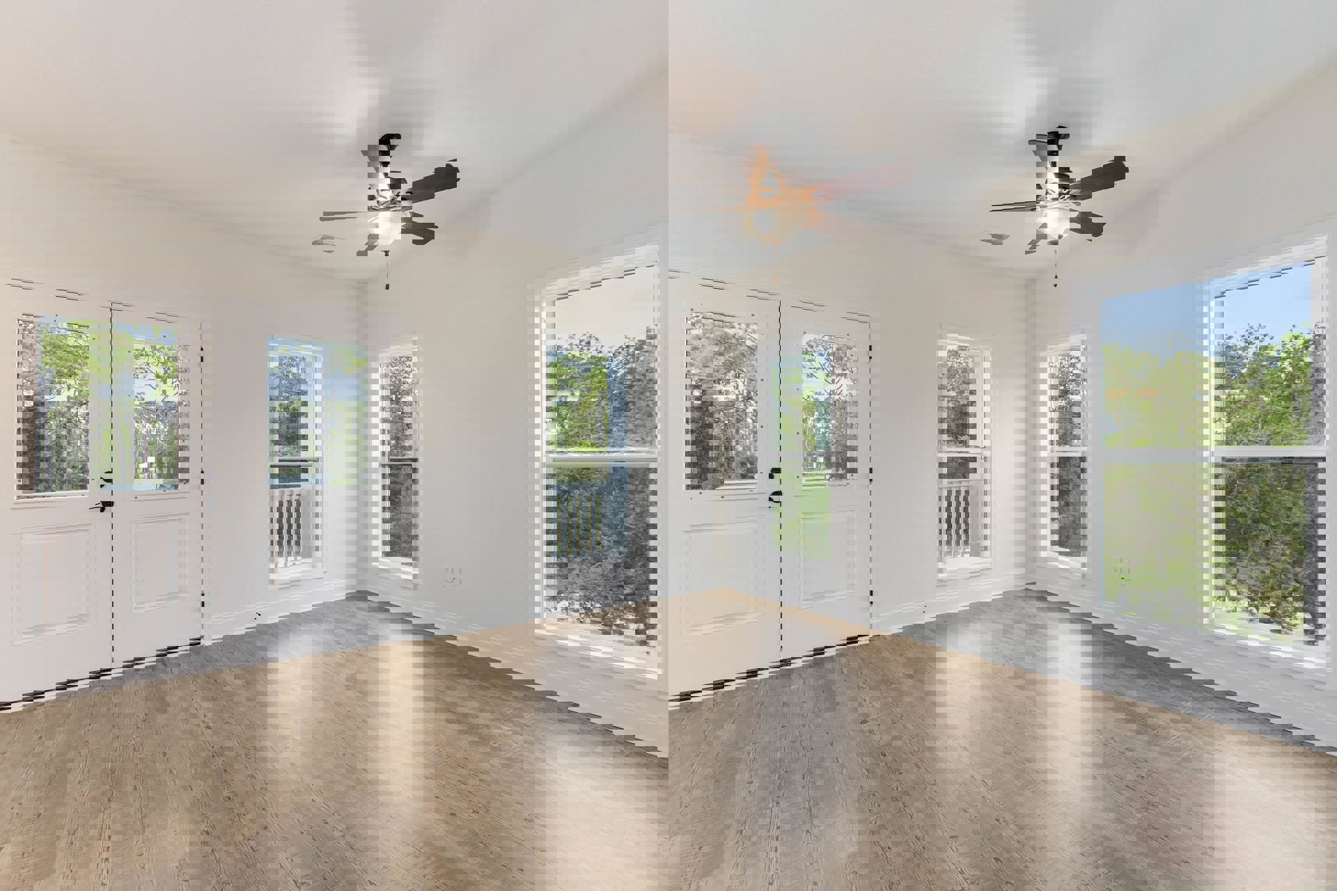 White-walled room with wood flooring, ceiling fan with light fixture, large window overlooking trees and forest.