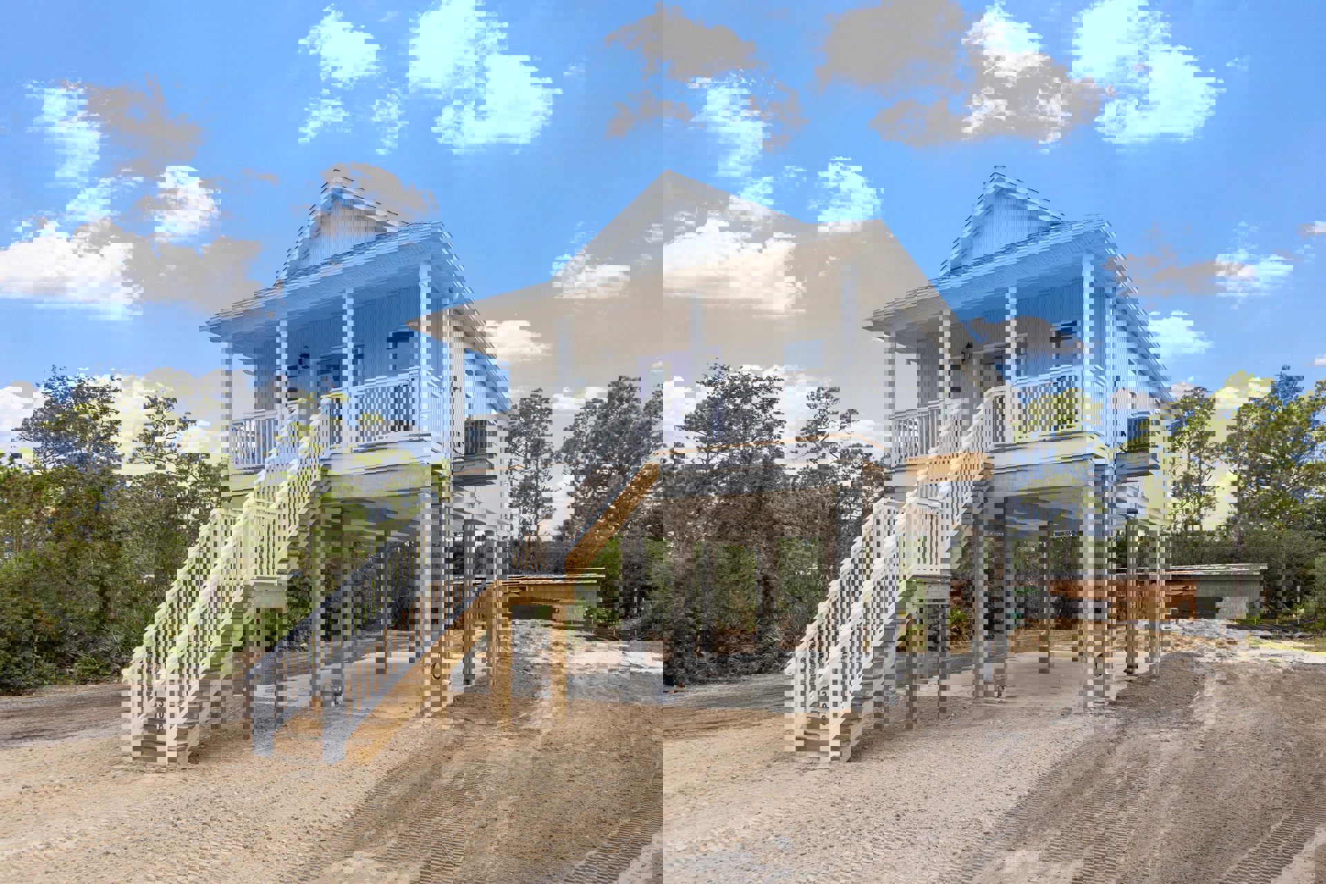 White exterior stairs leading to a porch, large window with white frame, dirt ground, blue sky, and trees surrounding a modern residential house