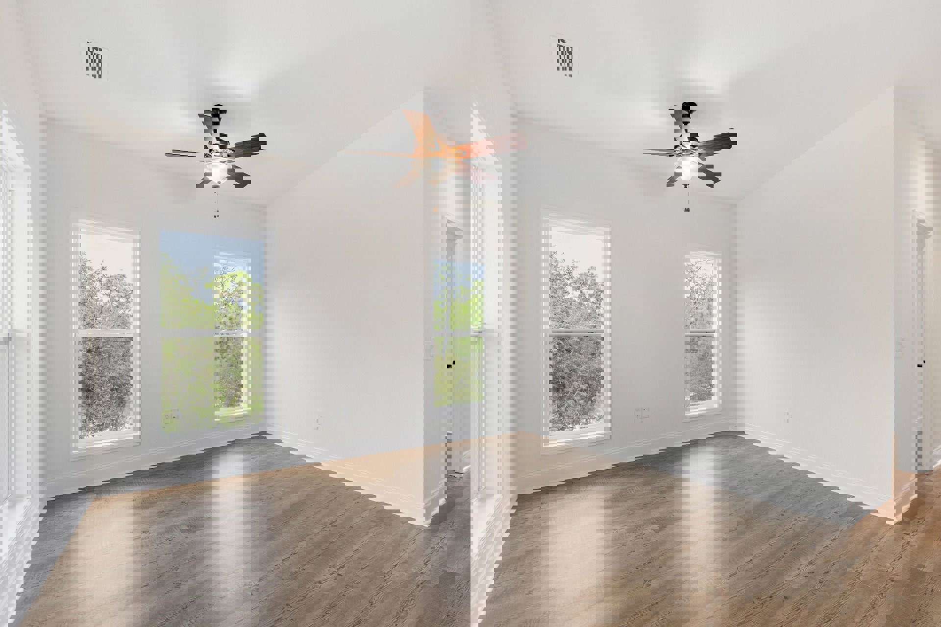 Ceiling fan with light fixture, large windows overlooking trees, wood laminate flooring, white plaster walls, and wall vent in a bright residential room