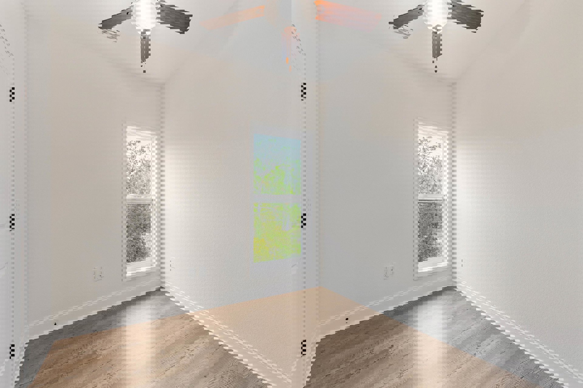 Ceiling fan with integrated light fixture above wood flooring, white walls, and large window framing forest view.