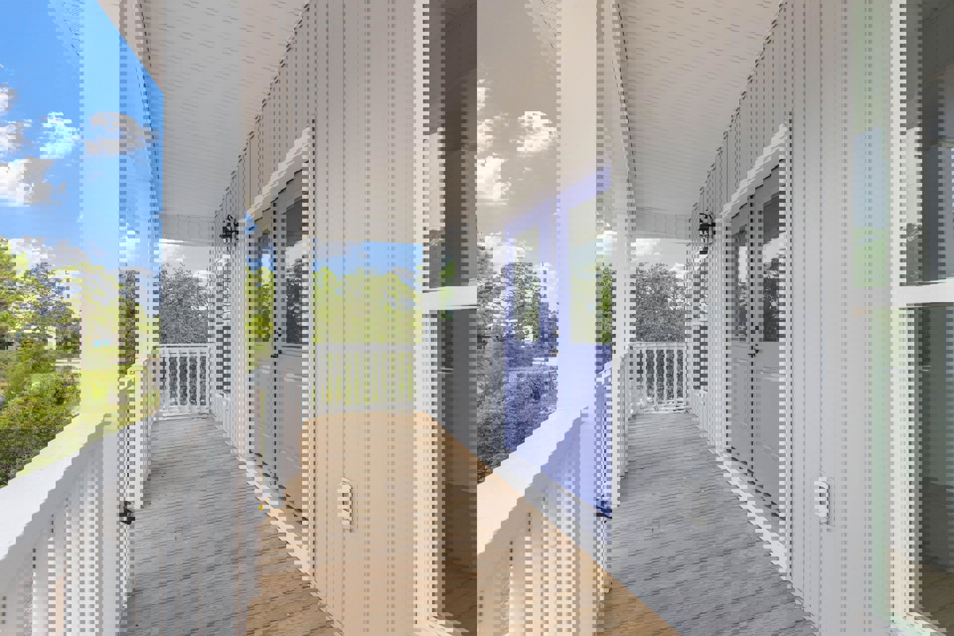 Blue front door framed by white porch railing, wooden deck flooring, window with white trim, outdoor lamp, leafy trees in background