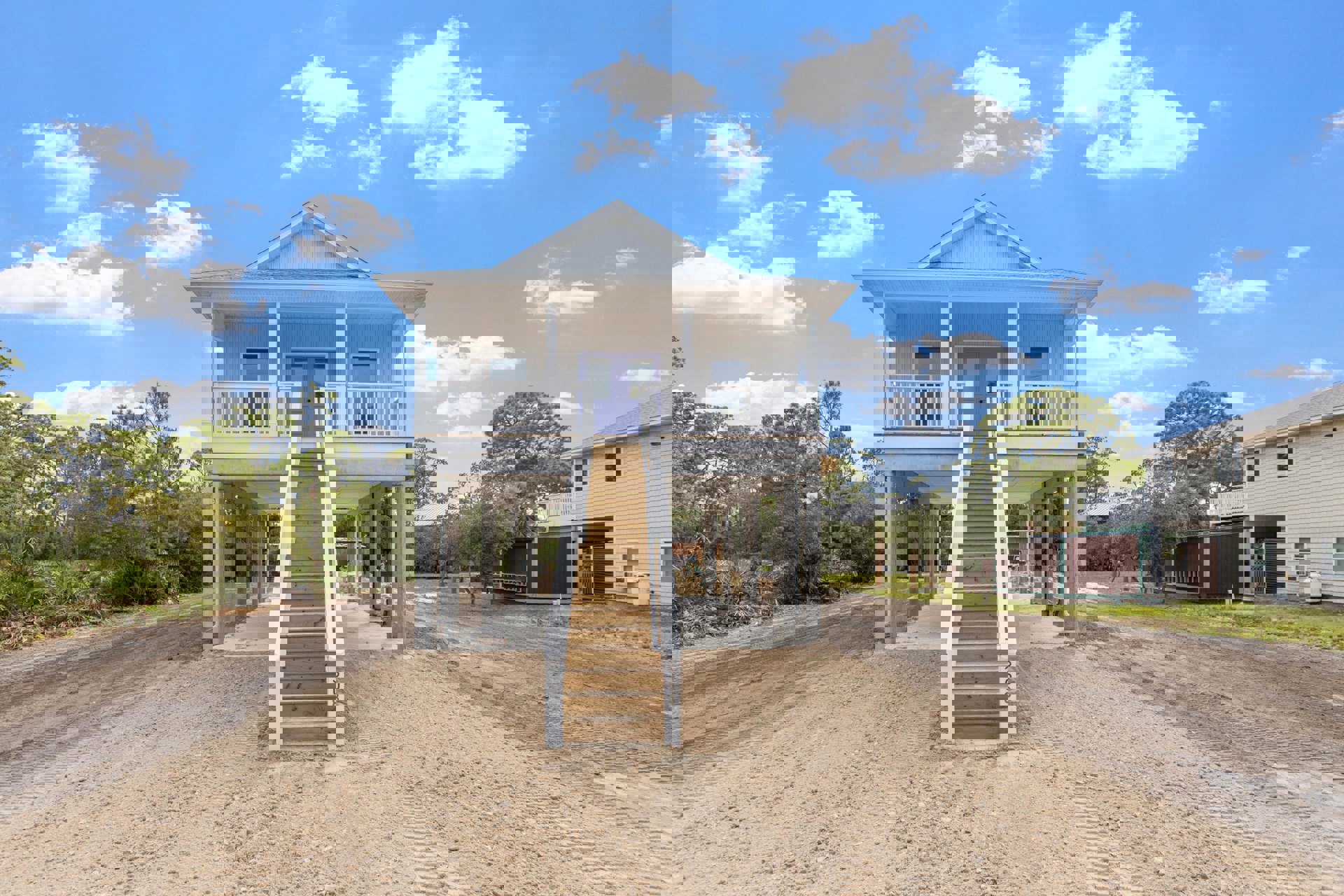 White porch with wooden stairs and white railings leading to a blue front door, surrounded by trees and plants under a cloudy sky.