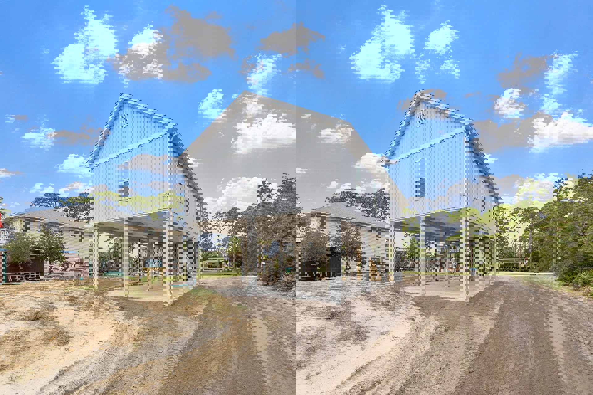 White custom home with expansive foundation, large roof, prominent windows, surrounded by dirt landscaping and trees under a blue sky with scattered clouds