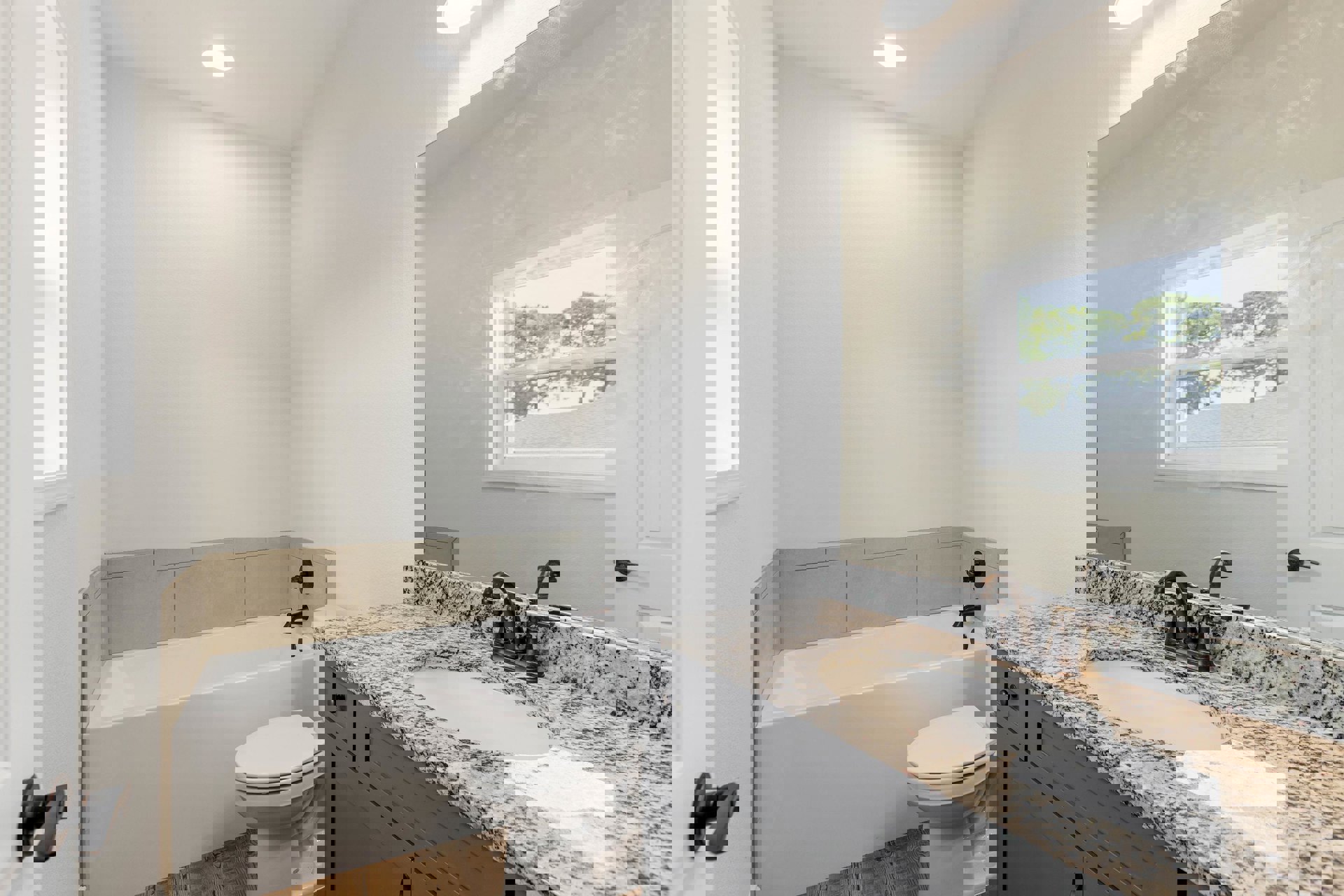 Marble countertop vanity with undermount sink and chrome faucet, freestanding bathtub, white tile flooring, toilet with closed lid, large window overlooking trees