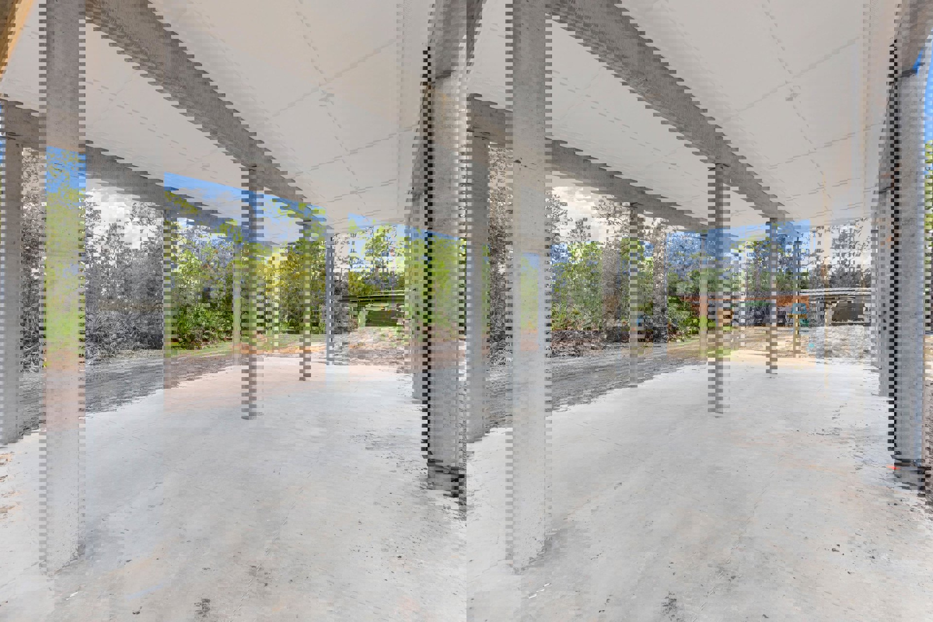 Concrete-floored exterior with tall pillars supporting a ceiling beam, surrounded by mature trees and shaded outdoor space