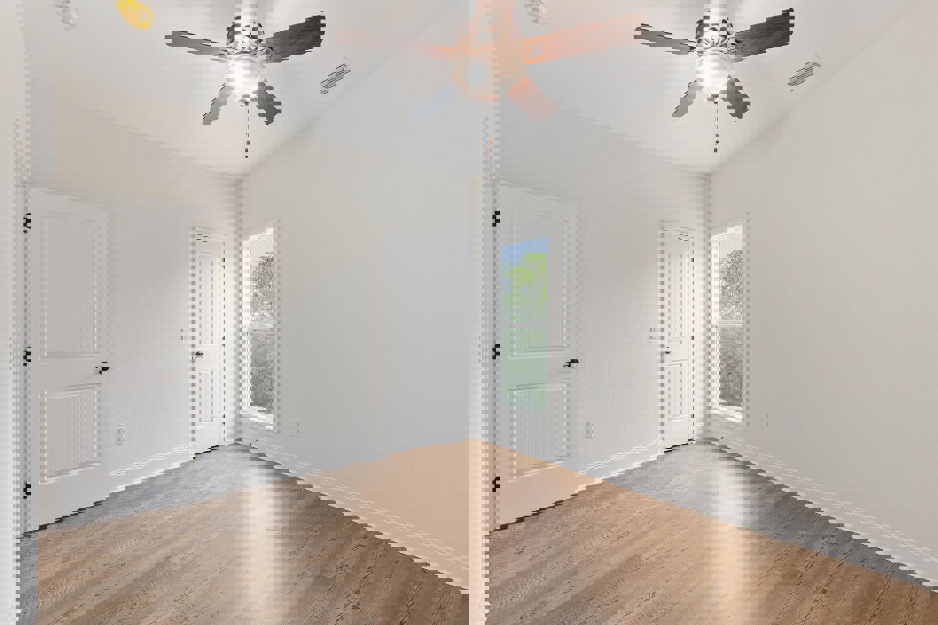 Ceiling fan with light fixture above wood flooring, two white doors with black handles, window showing trees outside, white walls and plaster ceiling