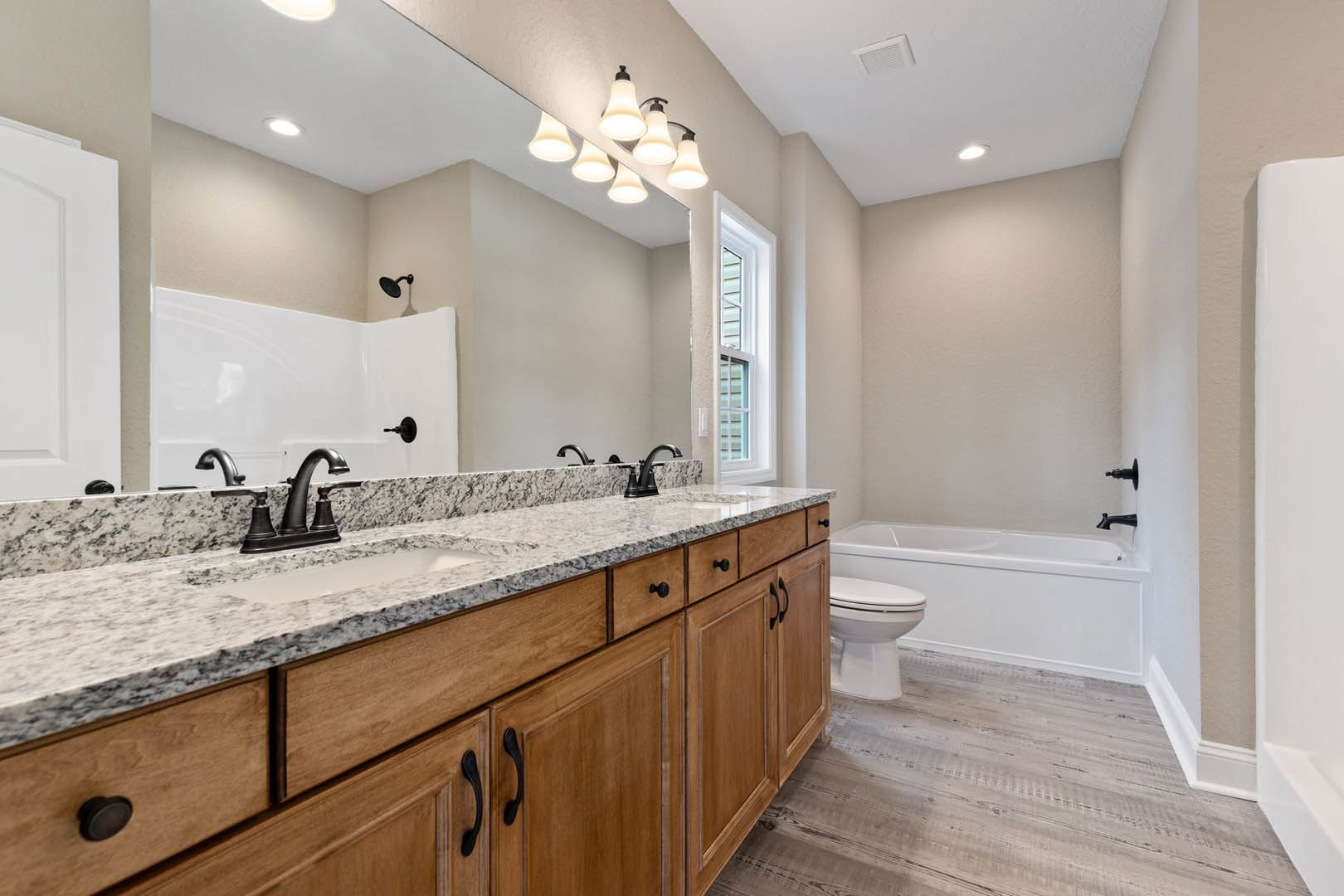 Bathroom with a wide mirror above a rectangular sink, white toilet, white bathtub with chrome faucets, light fixture with multiple bulbs, tiled walls, and cabinetry with drawers.