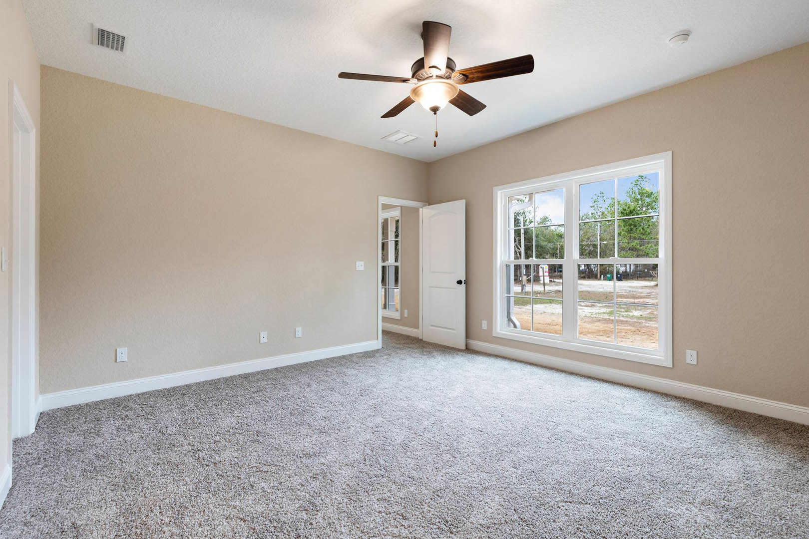 Carpeted room with white walls, ceiling fan with light, two windows overlooking yard, white doors with black hardware, crown molding along ceiling