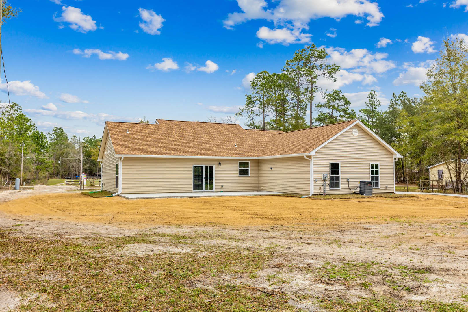 Modern home with light siding and dark roof, surrounded by a dirt yard and sparse grass, trees visible in the background, exterior door and heater unit near the side of the house