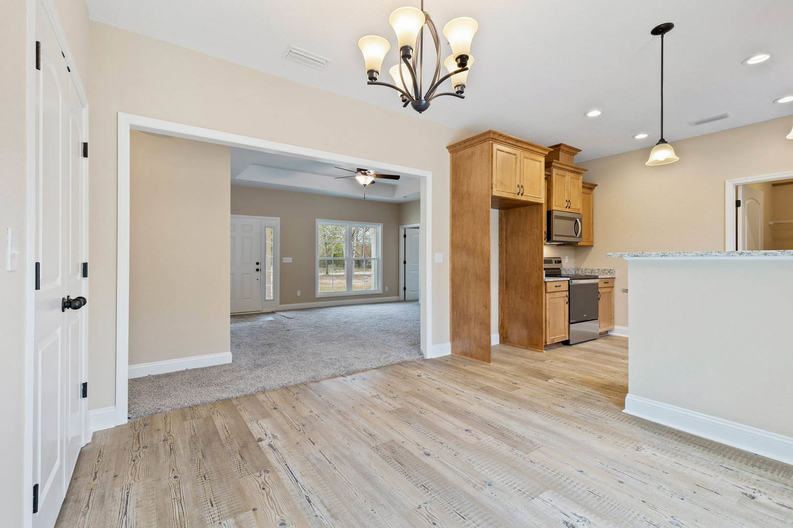 Open-concept living room and kitchen with hardwood floors, carpeted area, five-light ceiling fixture, white cabinetry, stainless steel microwave, large window overlooking trees