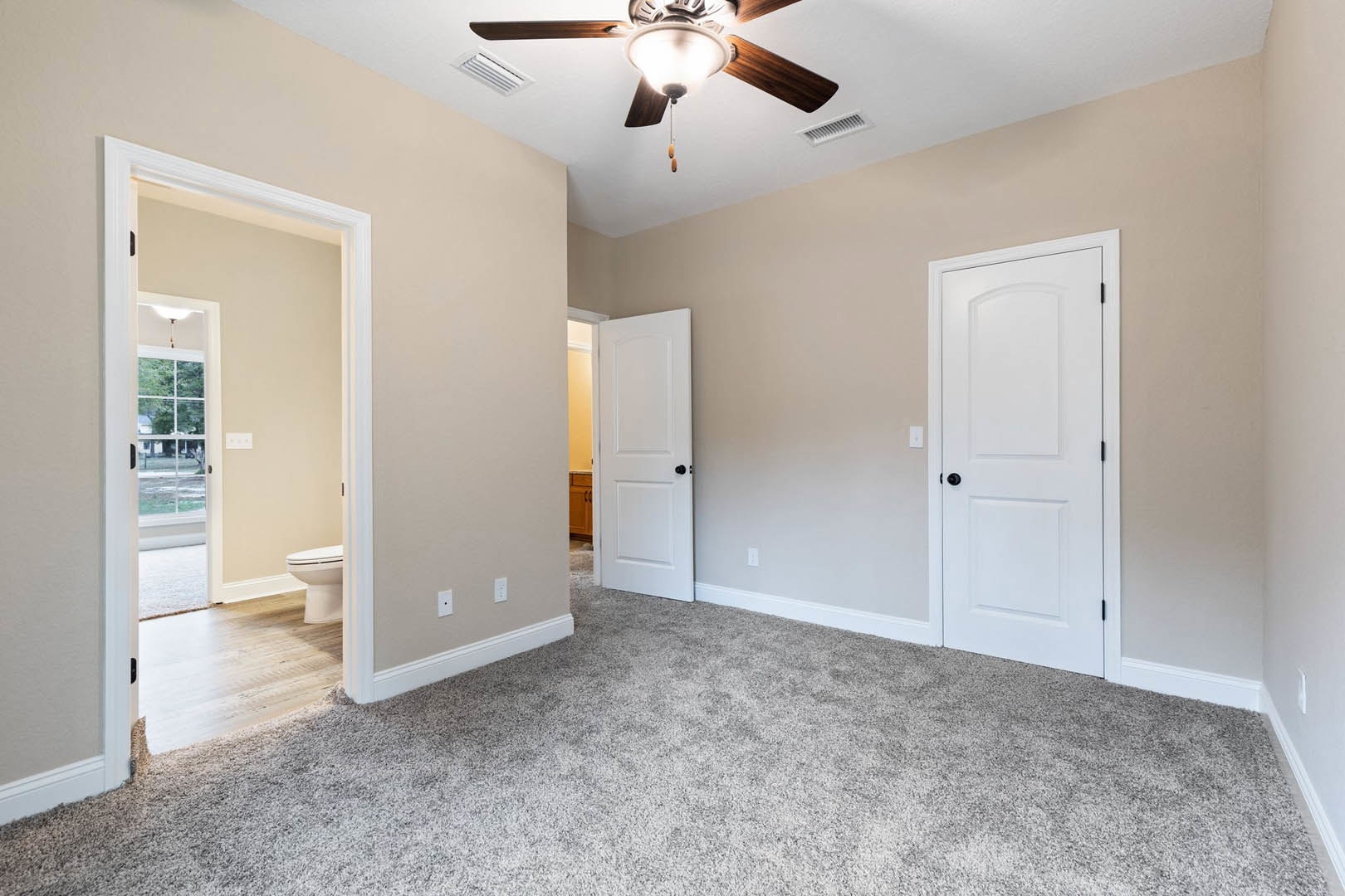 Carpeted bedroom with white walls, ceiling fan with light fixture, and white doors featuring black knobs; partial view of bathroom with toilet visible