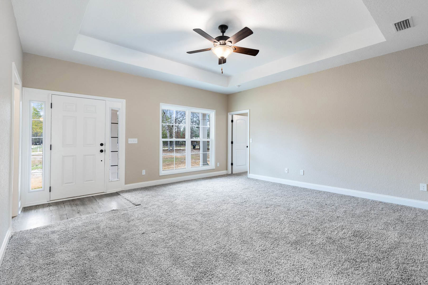 Carpeted room with white walls, ceiling fan with light, open white door with black handles, wall vent, large window showing trees outside