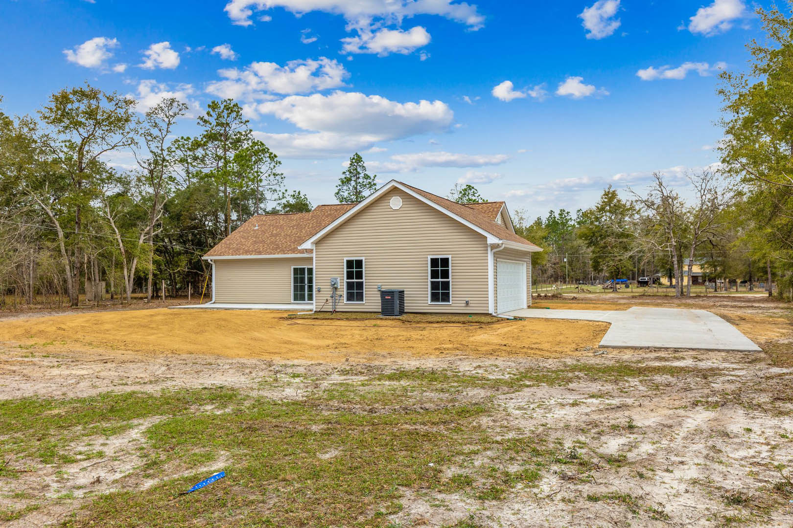 Two-story house with light siding, white-framed windows, and asphalt driveway; mature trees and grass lawn in background, dirt patch near entrance, blue object and black metal item