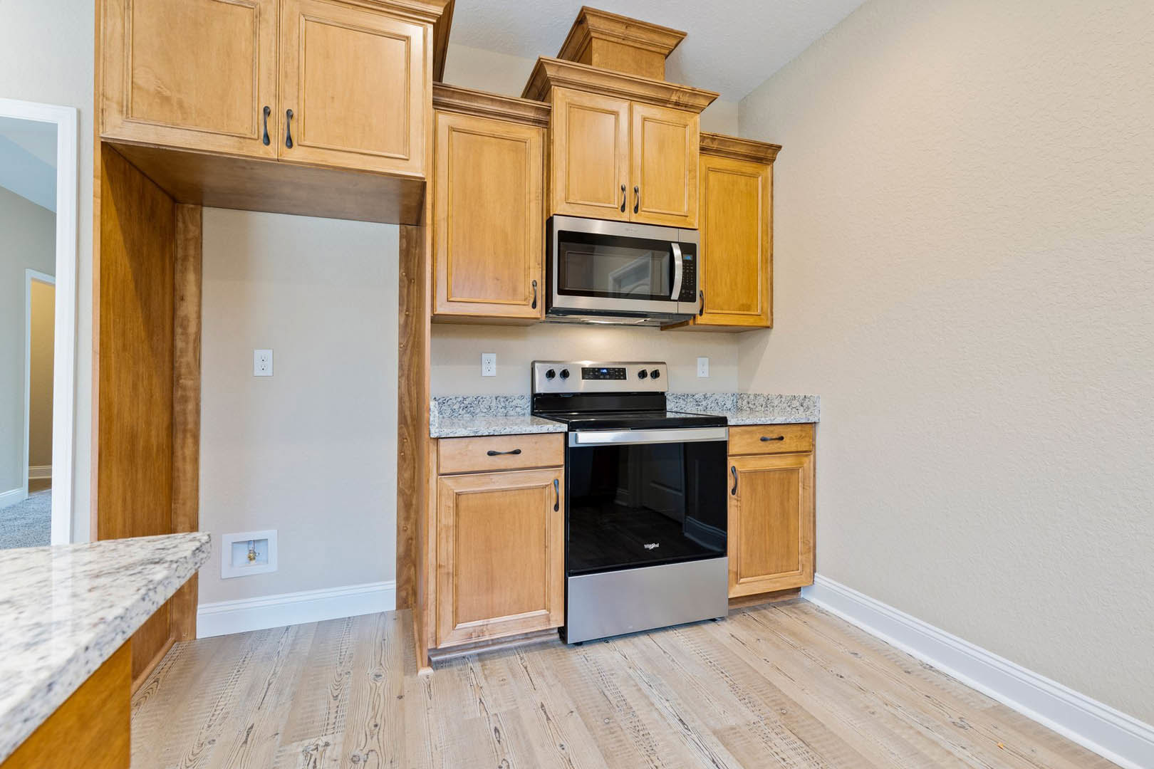 Kitchen with natural wood cabinets, stainless steel microwave, light-colored countertop, built-in dishwasher, white wall with light switch, and wooden door.