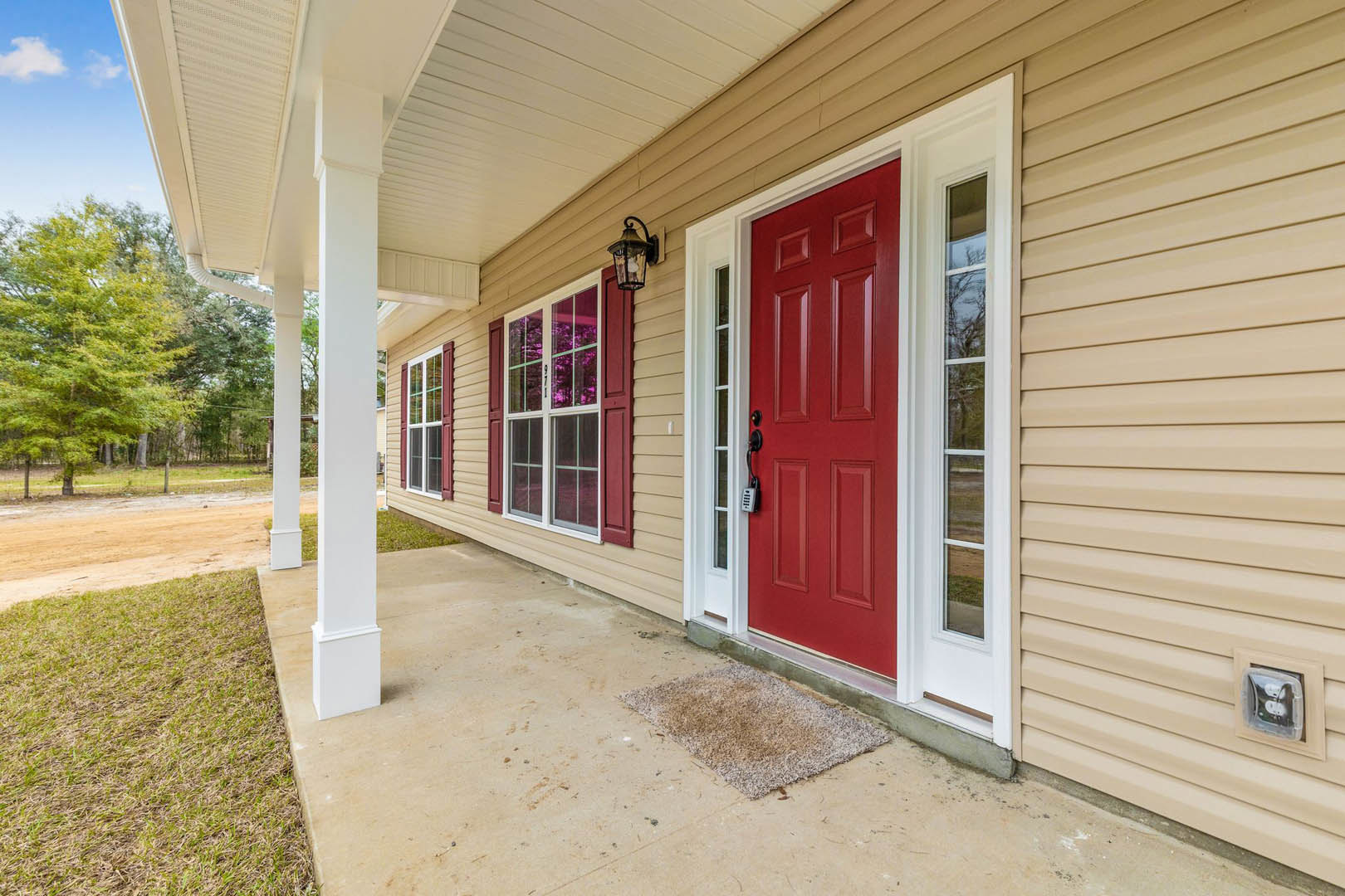 Red front door with silver lock, flanked by white pillars on a porch, white siding exterior, green tree and grass in foreground, blue sky above.