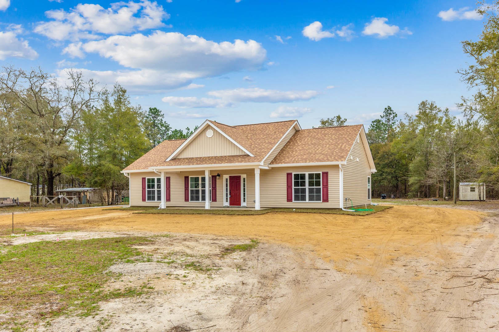 Two-story house with red doors and white-trimmed windows, surrounded by trees, dirt road and patch in front, under a blue sky with scattered clouds