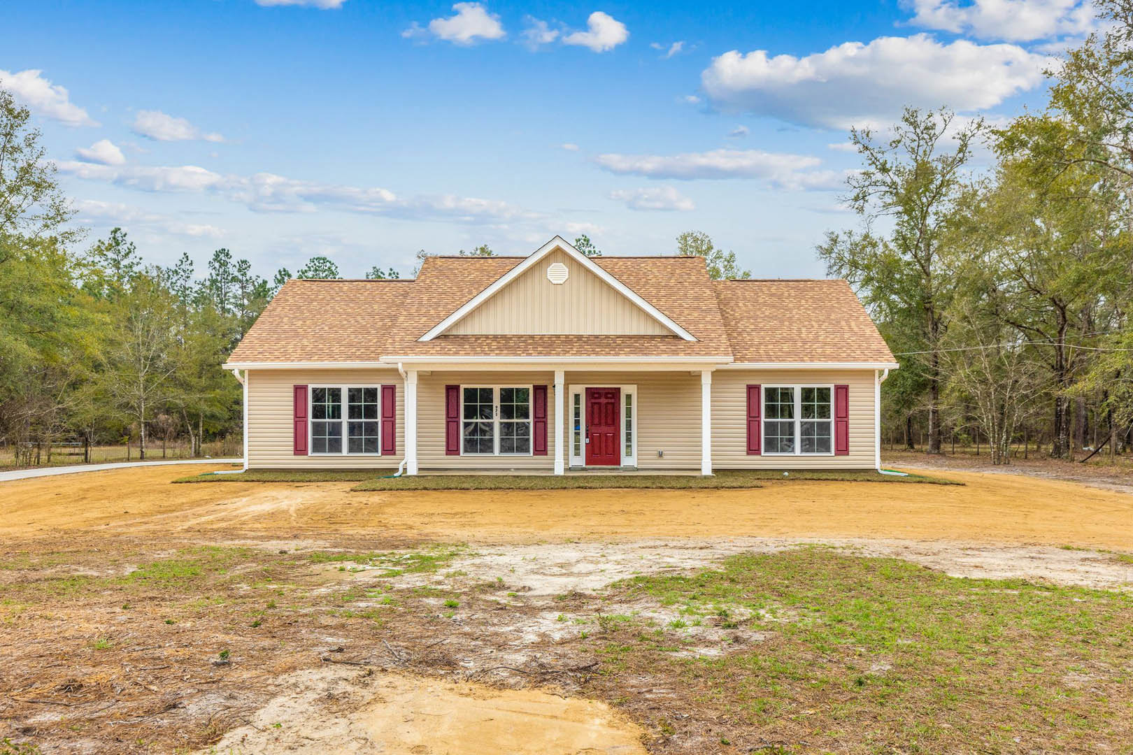 Two-story house with white siding, red front door, white-framed windows, and a dirt yard in front, surrounded by sparse grass and trees under a cloudy sky