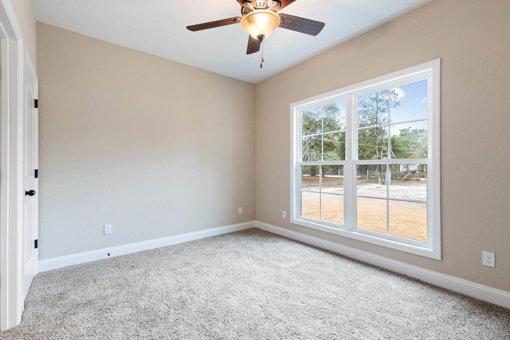 Neutral-toned room with white carpet, ceiling fan with light fixture, large window showing trees outside, white electrical outlet on wall, simple plaster walls and molding.