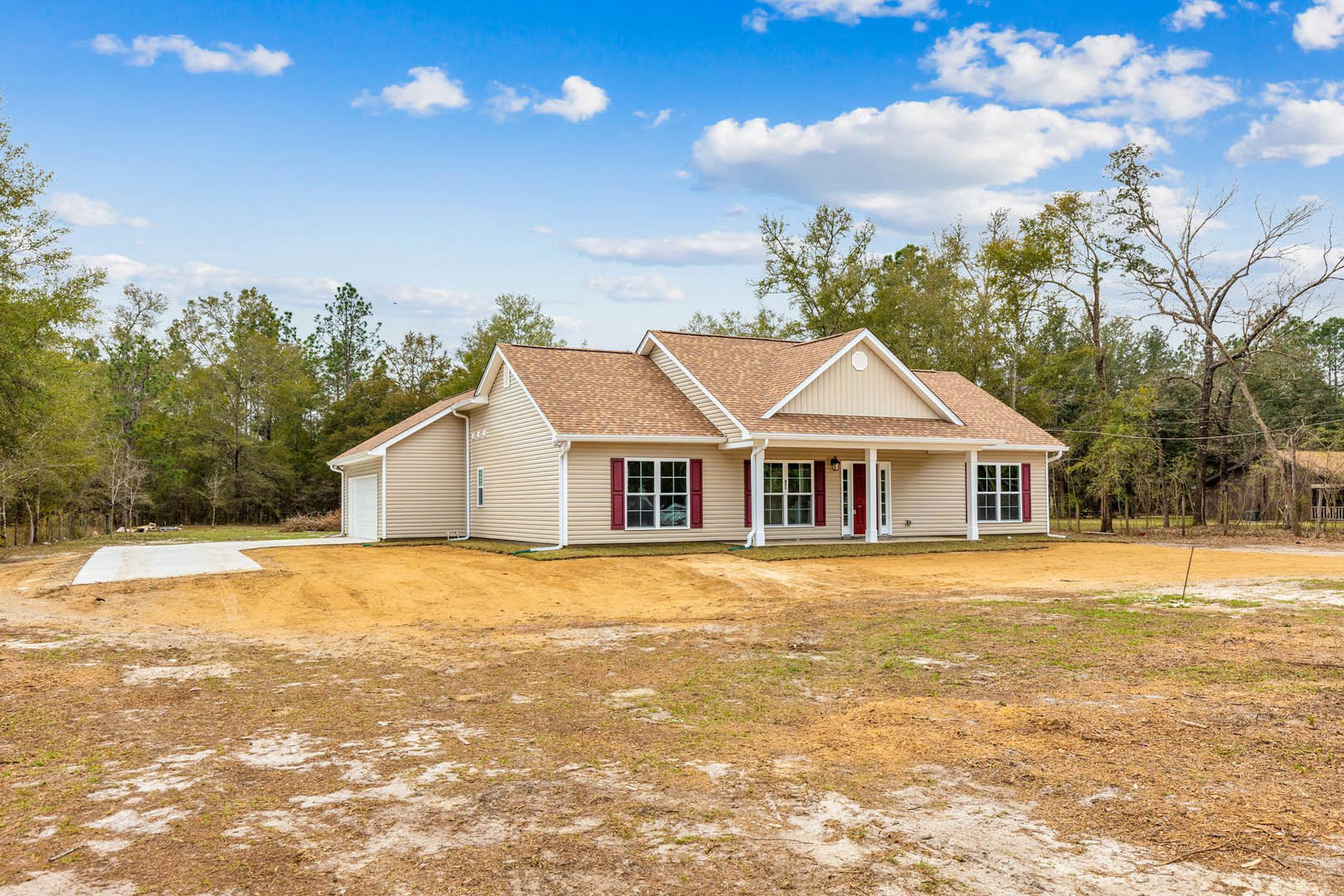 Two-story house with red shutters, white door with glass panes, dirt yard in front, trees and blue sky with clouds in background