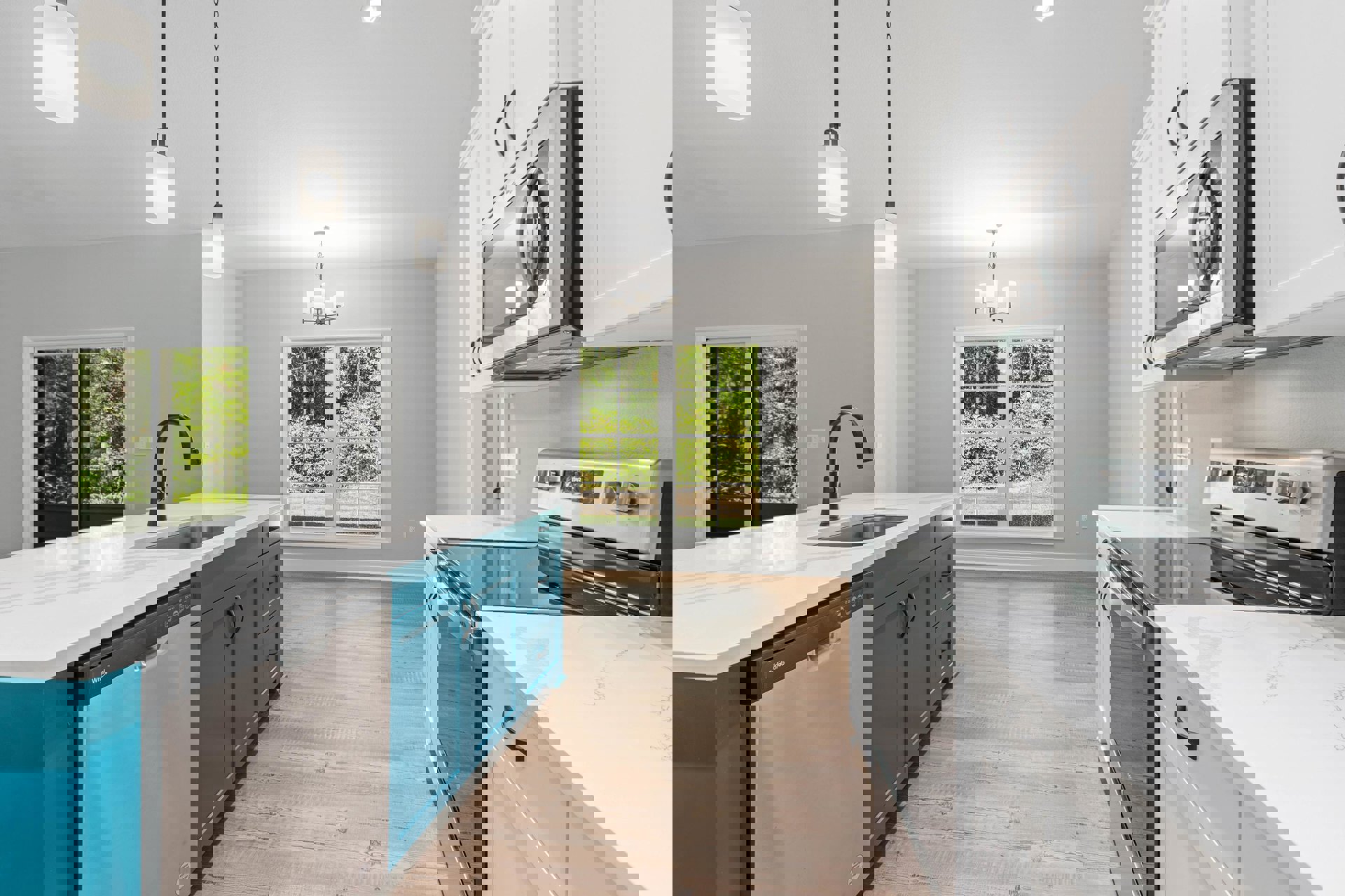 Kitchen with white upper cabinets and blue lower cabinets, stainless steel stove and microwave, quartz countertops, pendant light fixture illuminated, window overlooking trees