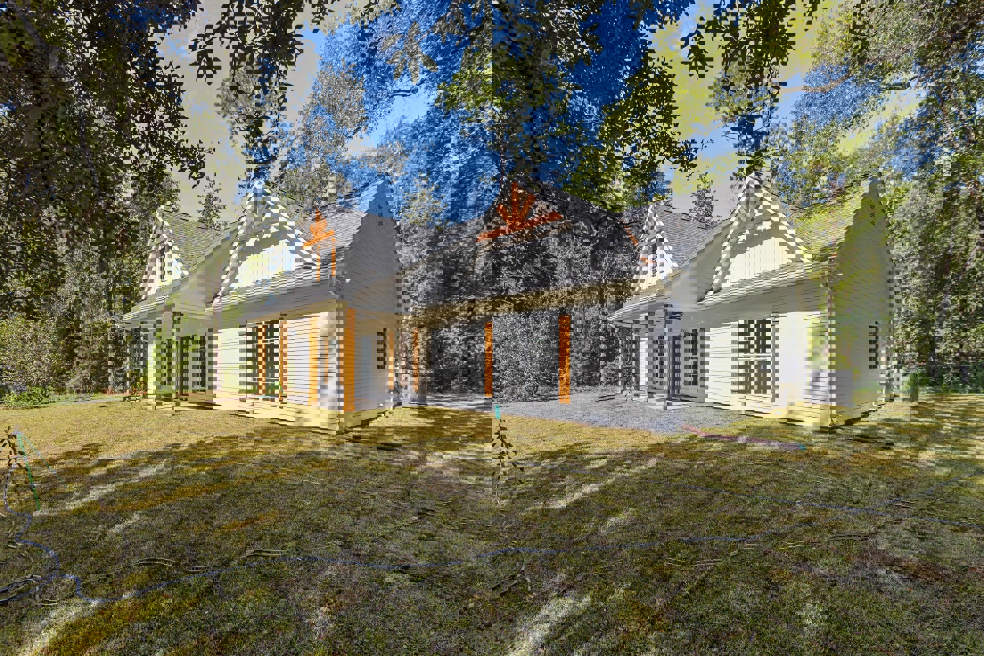 White siding home with gabled roof, large front window, green lawn, garden hose, and mature trees in the background