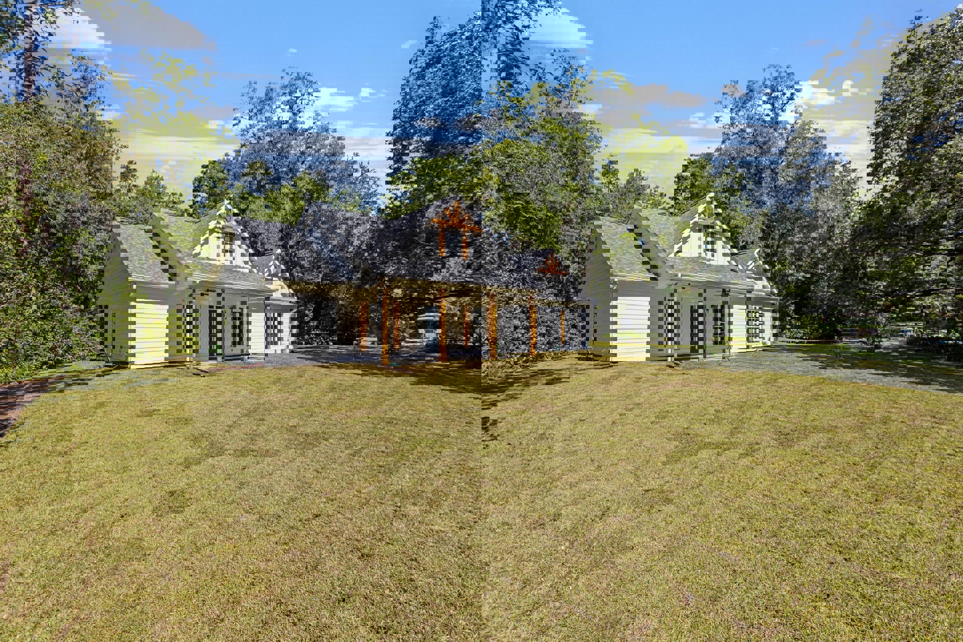 Two-story farmhouse with white siding, blue front door, covered porch, attached garage, expansive green lawn, mature trees in the background, partly cloudy sky overhead