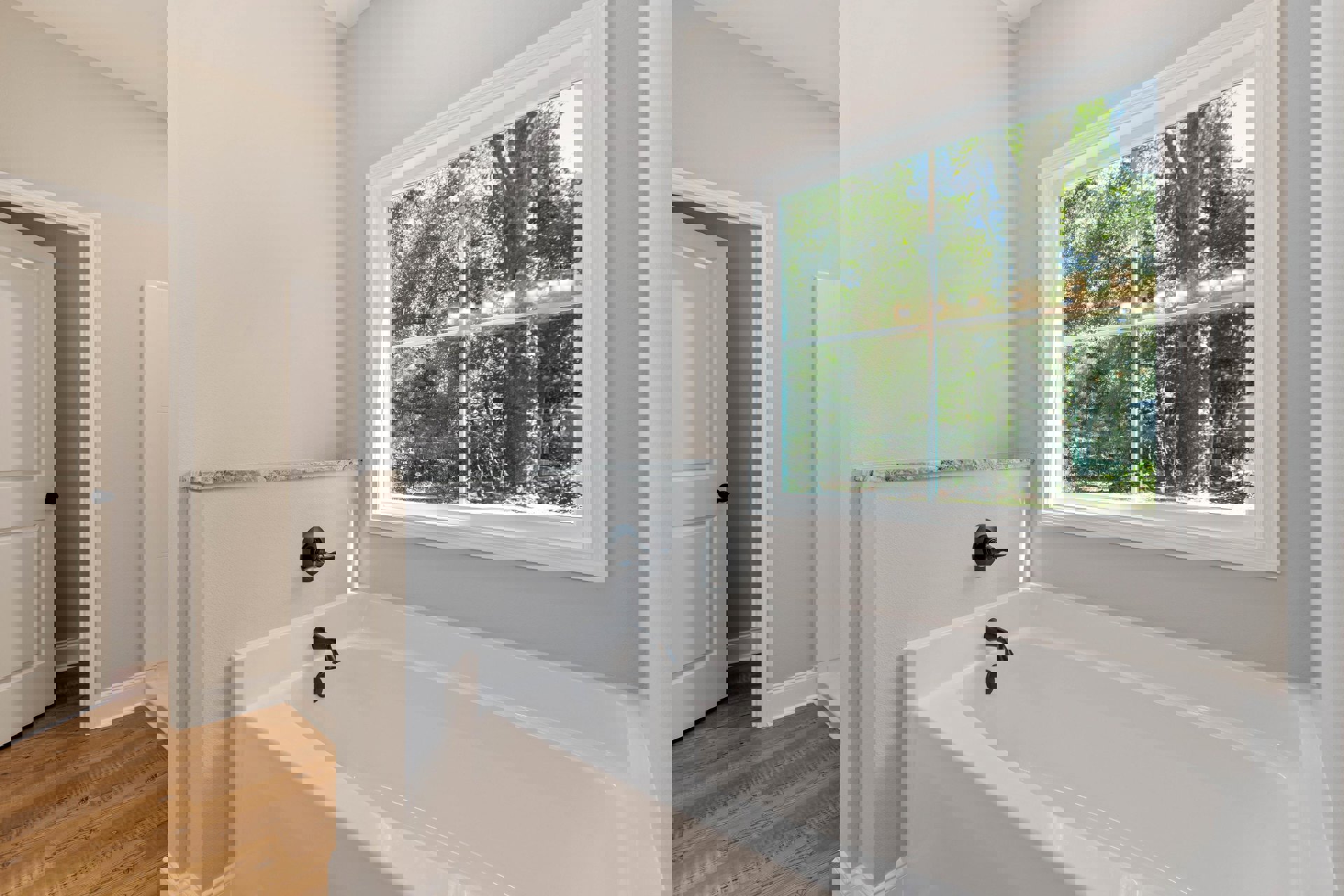 Freestanding white bathtub with chrome handle on wood floor, white walls, black wall knob, large window overlooking trees