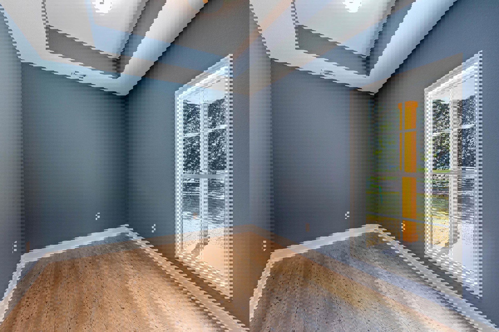 Sunlit room featuring a large window framed by a wood post, tree visible outside, wood flooring with white baseboards, white plaster walls, and ceiling-mounted light fixture.