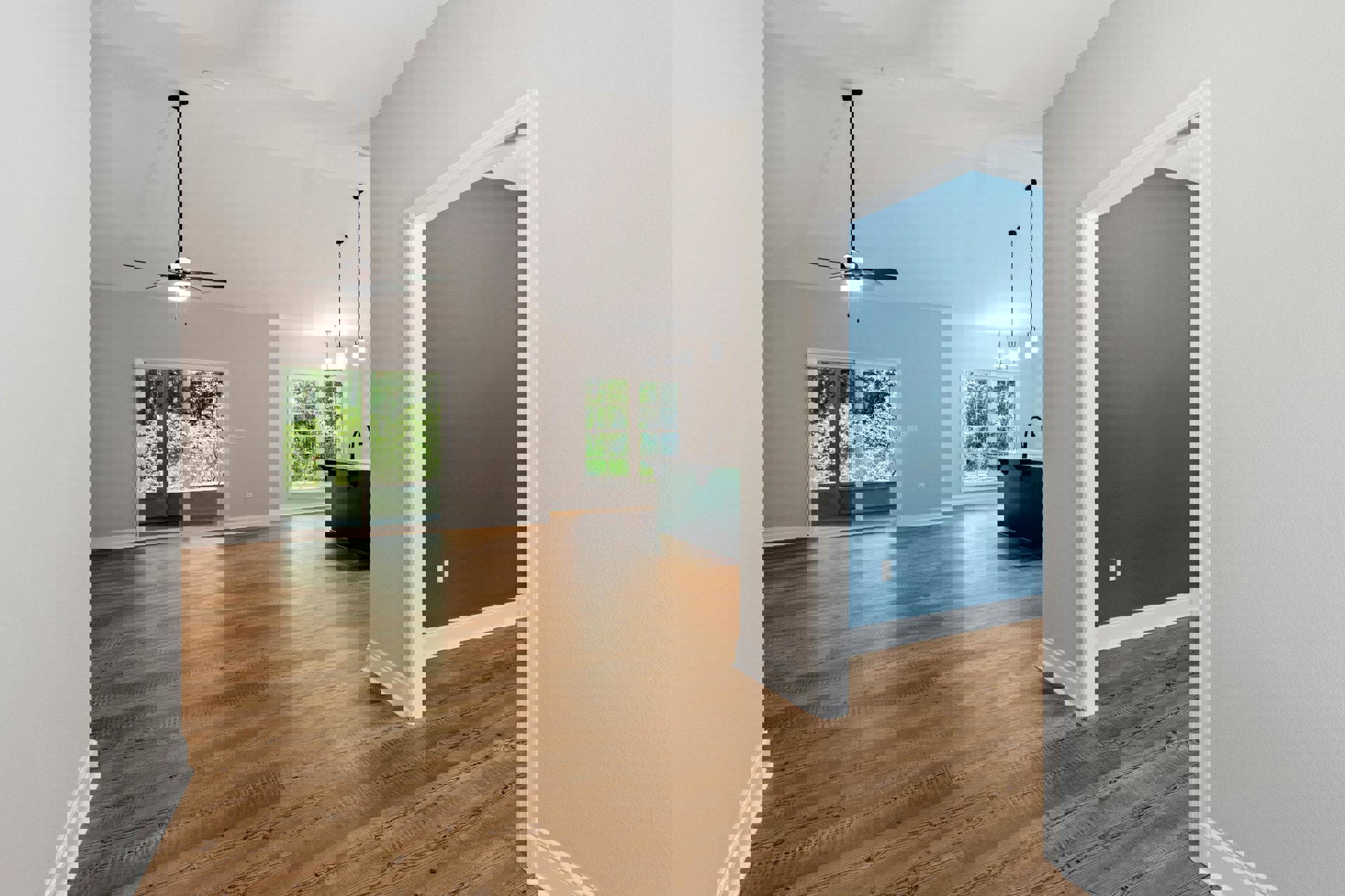 Wood flooring and white walls in a room with a black door, blue accent wall with white trim, ceiling fan, large window overlooking trees, green countertop with black appliance.