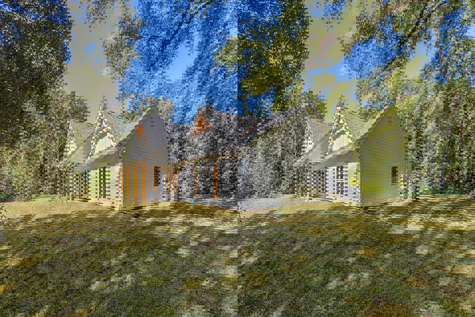White house with glass-paned windows, grey roof and gutter, surrounded by mature trees and green lawn with garden hose; rectangular vent visible on exterior.