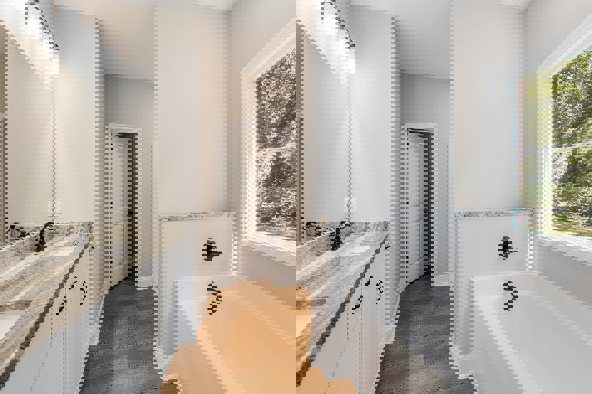 Freestanding white bathtub with chrome faucet beside a large window overlooking trees, modern sink with black hardware, white door with black knobs, light tile flooring and walls