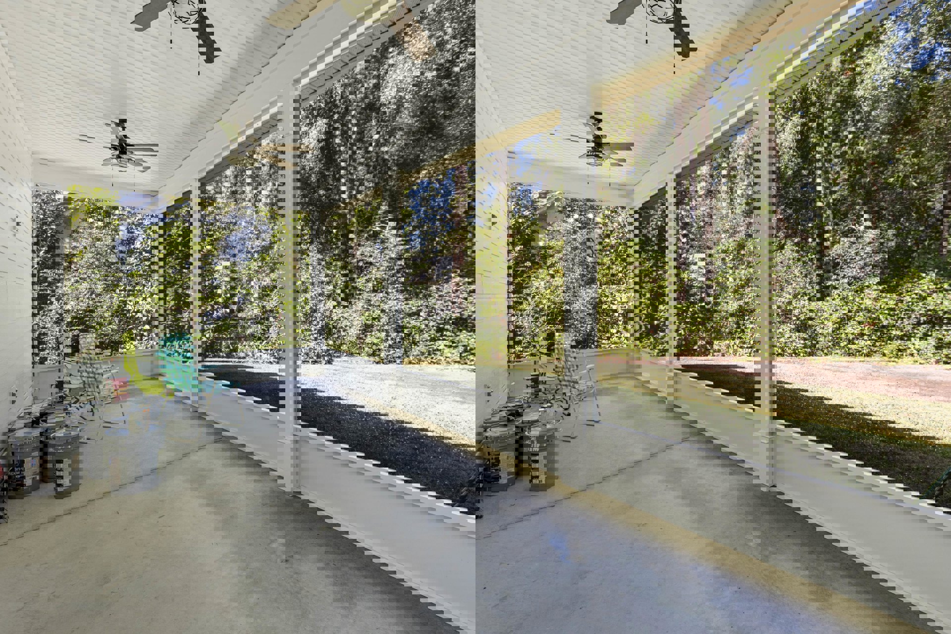 Covered porch with concrete floor, white wall, windows, ceiling fan with light, single lawn chair, paint buckets, shaded by nearby trees and plants