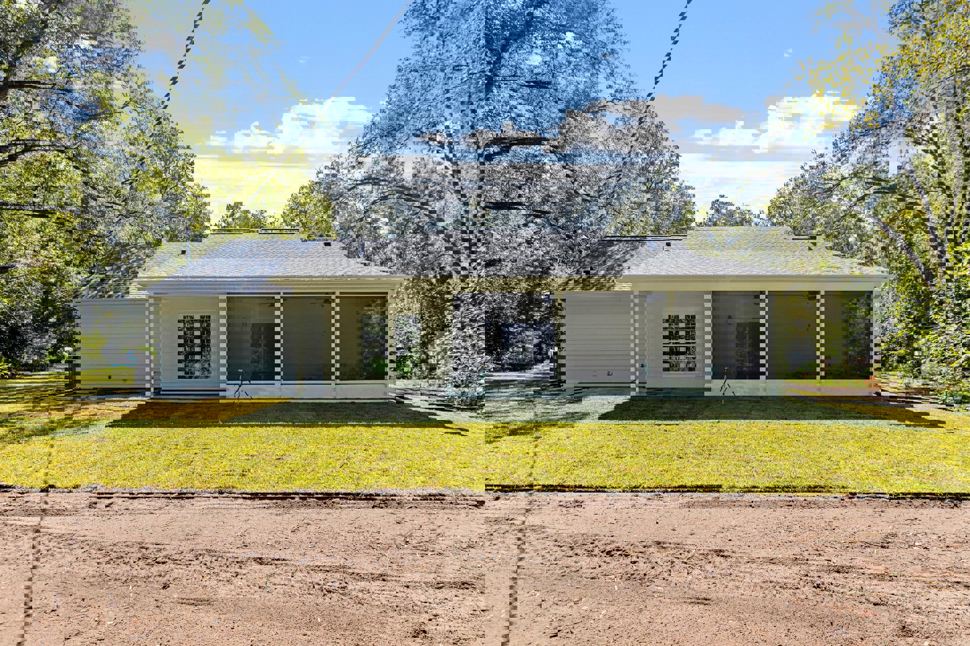 Two-story home with multi-pane windows, sliding glass door, dirt road with tire tracks, green lawn, mature trees, tripod and hose in foreground