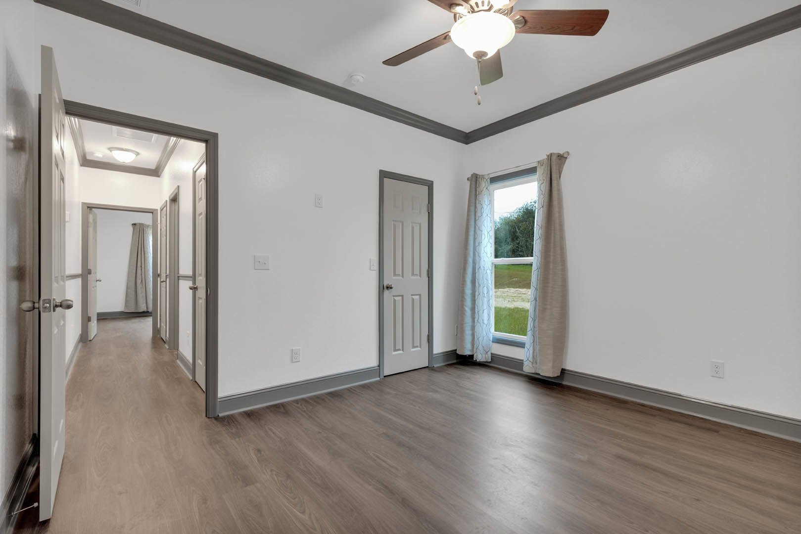 Bedroom with wood flooring, white door, ceiling fan with light fixture, and window overlooking grassy field