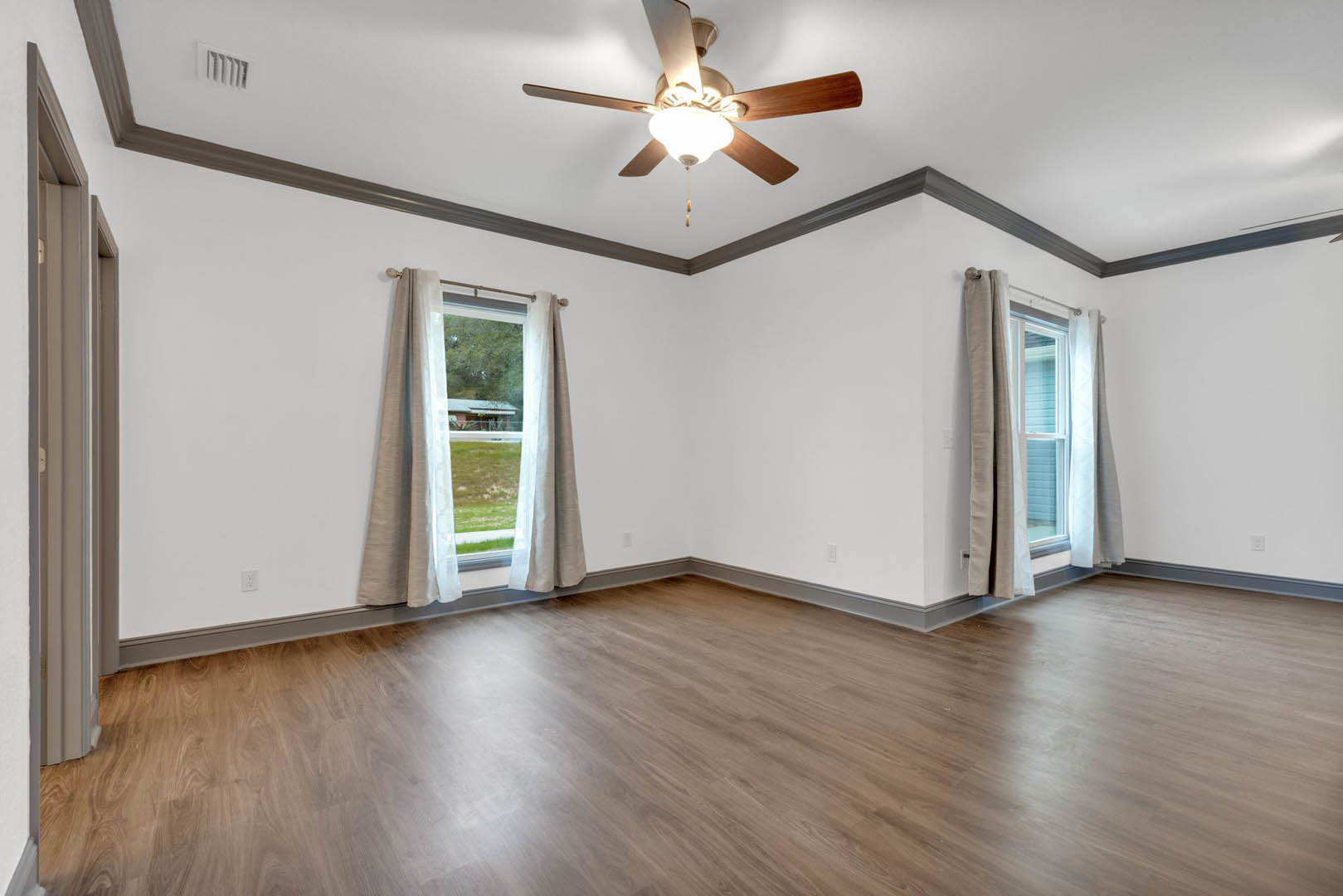 Ceiling fan with light fixture above hardwood floor, large windows with white curtains, plaster walls, view of neighboring house through window