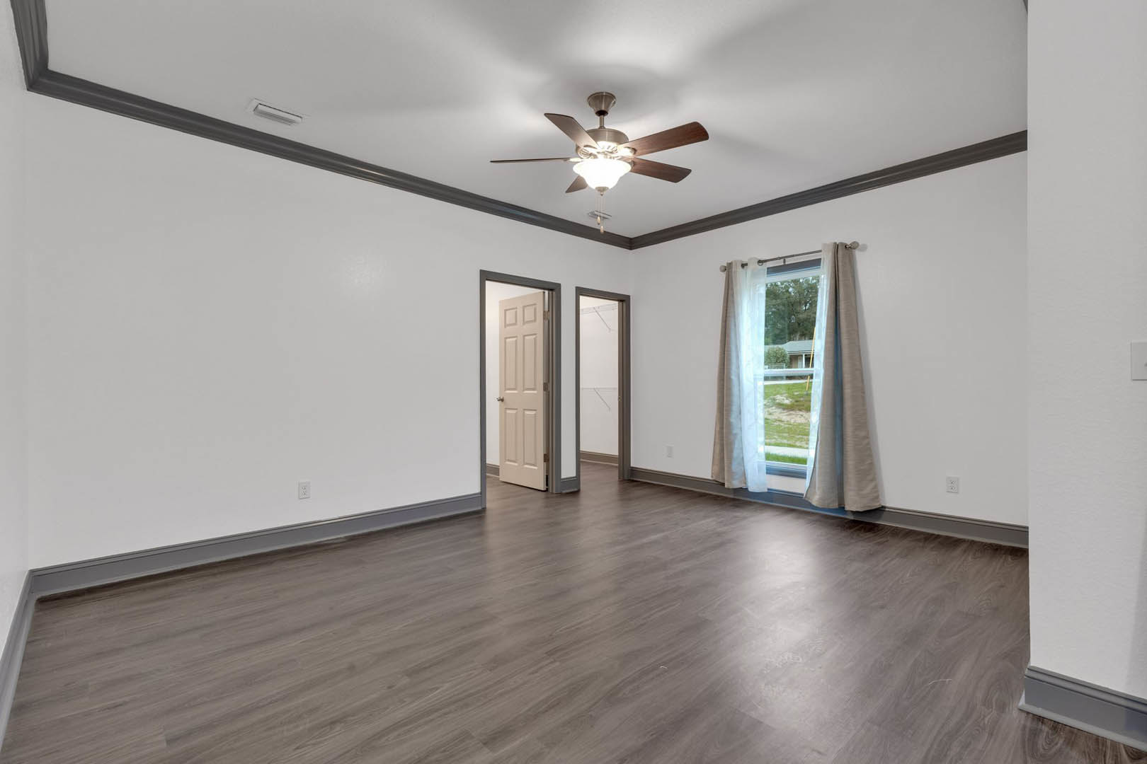 Ceiling fan with light fixture above wood laminate flooring, white plaster walls, open white door with silver handle, window showing exterior house view