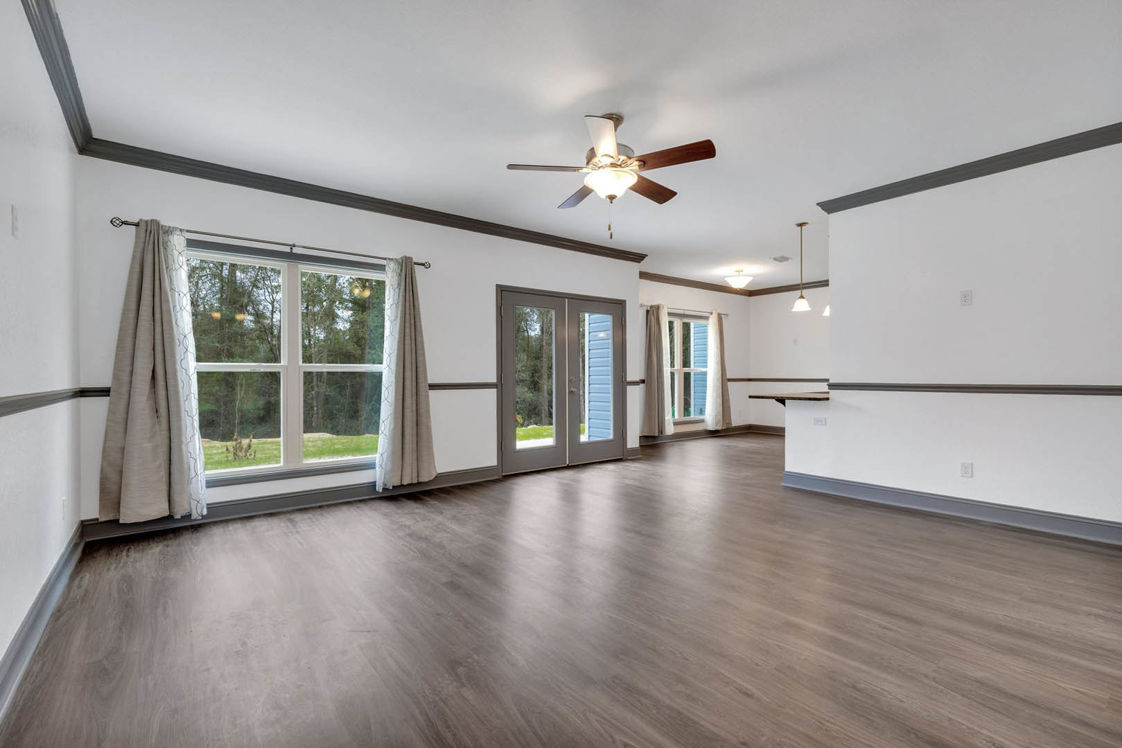 Living room with wood flooring, ceiling fan with light fixture, double glass doors, and window dressed with curtains overlooking trees.