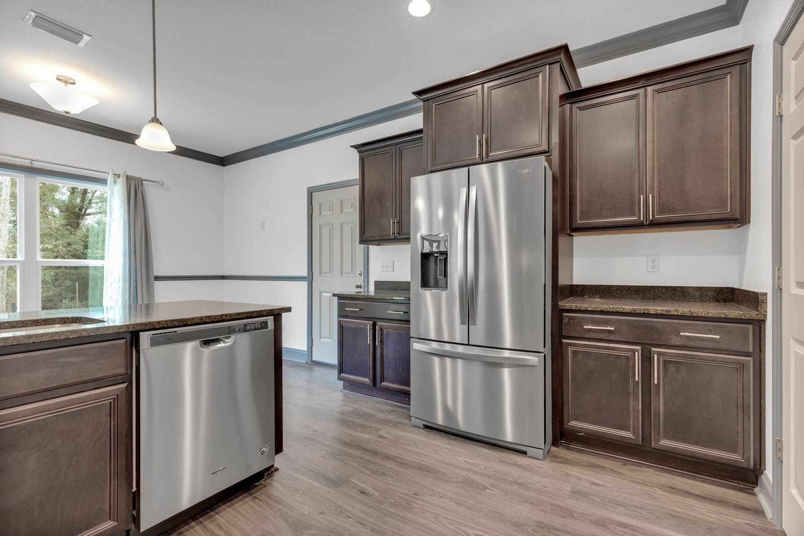 Modern kitchen featuring stainless steel refrigerator, white cabinetry, stone countertops, and a window with sheer curtains overlooking leafy trees