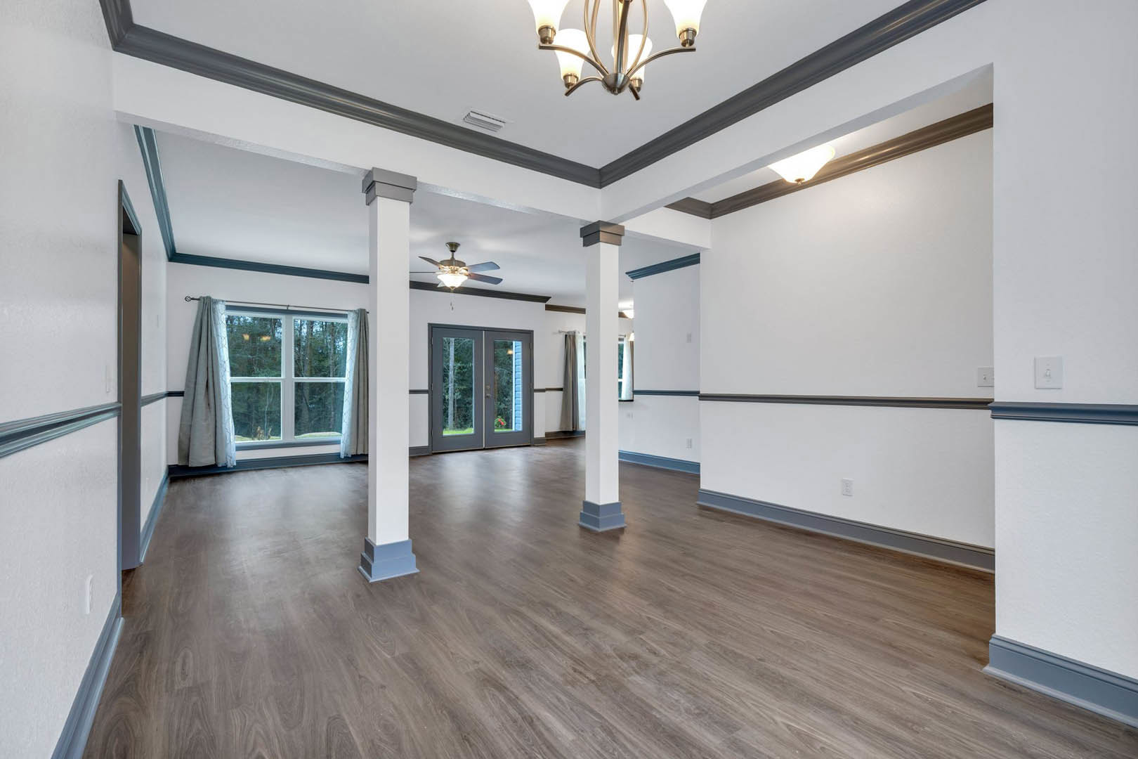 Wood floor room with white pillars, double glass doors, ceiling fan, chandelier, and window with sheer curtain overlooking trees.