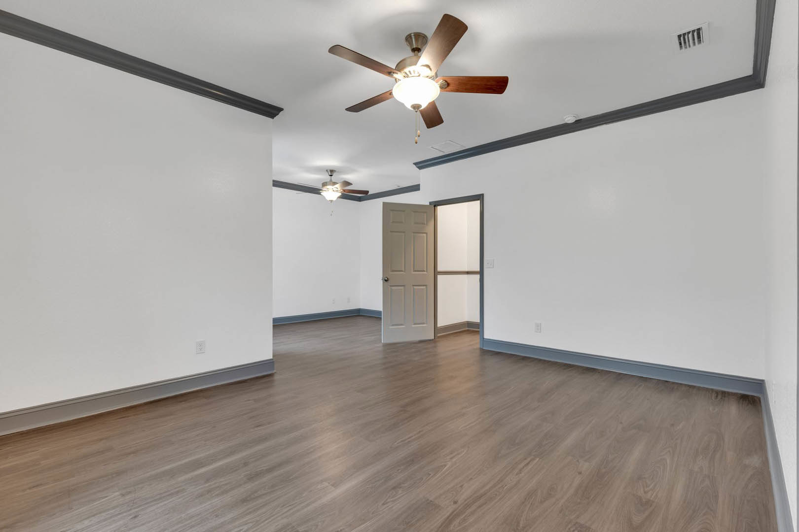 Ceiling fan with light fixture mounted above wood flooring, white walls, white door with silver knob, wall vent visible