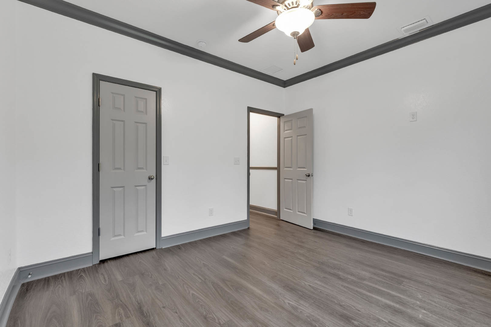 Wood flooring and white walls in a room with a ceiling fan and light, white doors with silver knobs, and a brown bar along one wall
