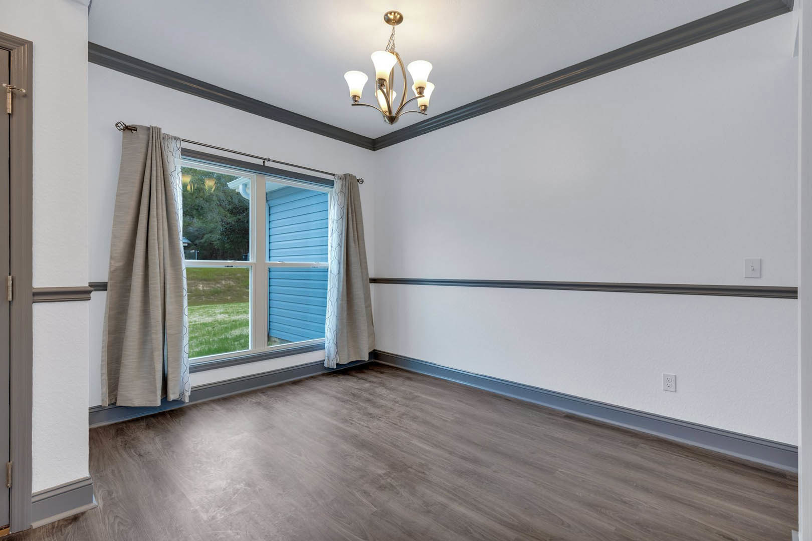 Wood floor with white walls and blue trim, large window framed by curtains, chandelier overhead, and decorative molding along ceiling.
