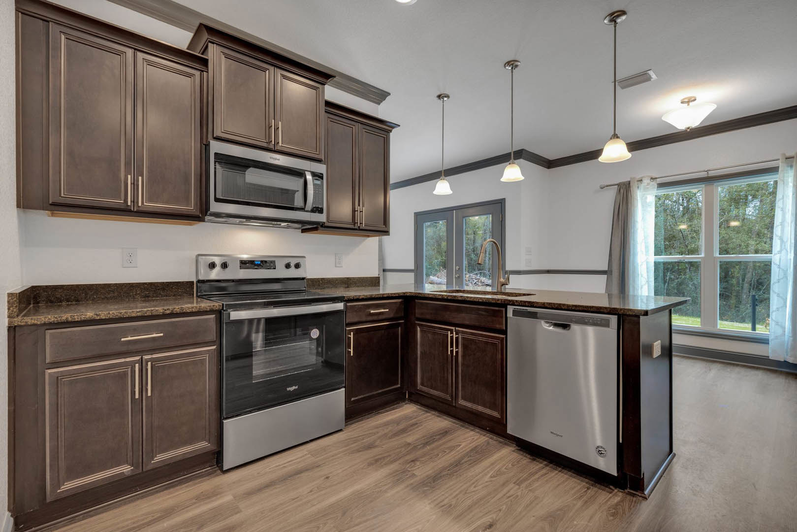 Kitchen with dark wood cabinets, stainless steel oven, microwave, and dishwasher, light stone countertops, window above sink, and dining table adjacent to counter.