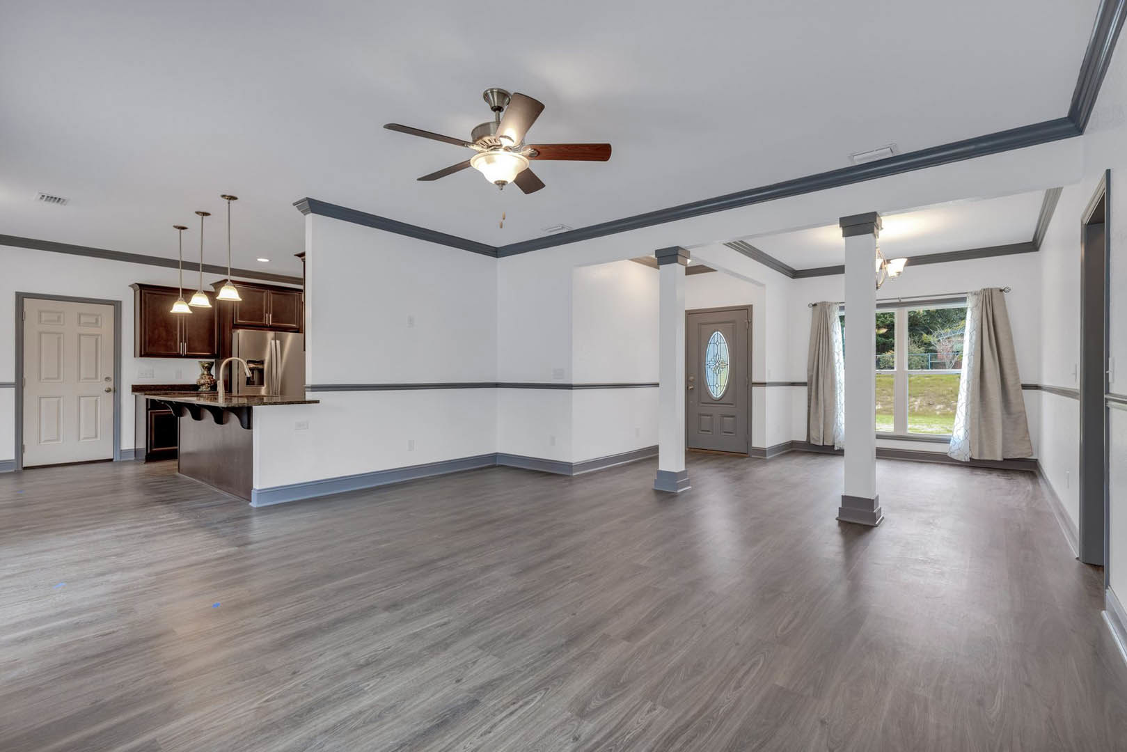 Living room with wood flooring, white walls, ceiling fan with light fixture, white door with silver handle, grey curtain, and plaster ceiling