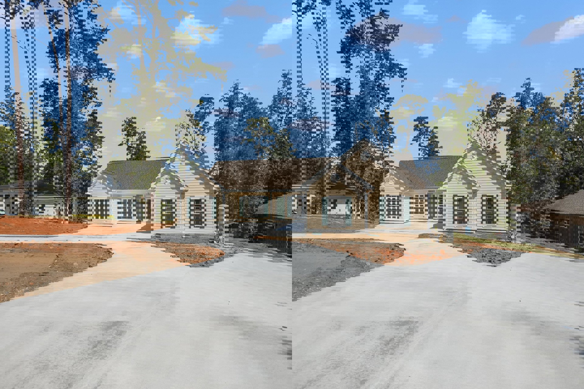 Two-story house with green front door, green shuttered windows, paved driveway, large tree in front yard, metal cylinder with white sign near porch, trees and cloudy sky in