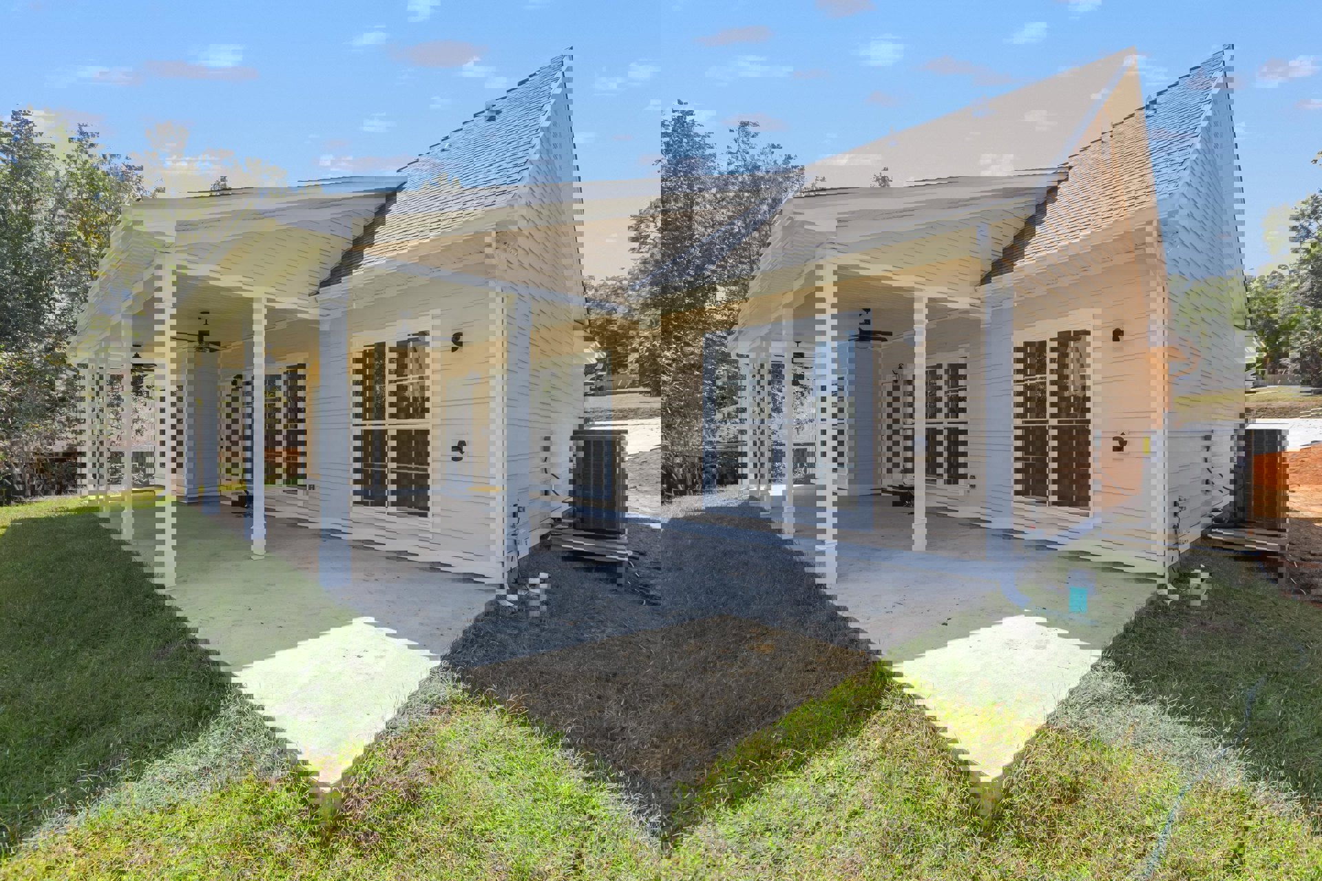 Modern home exterior with covered porch, white-framed windows, large grey heat pump beside concrete patio, green lawn, and surrounding trees