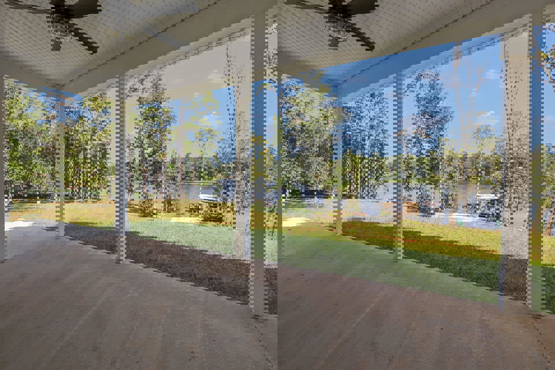 Covered porch with white pillars and siding, overlooking grassy yard, lake, trees, boat dock, and bridge with parked truck; close-up of outdoor lamp in foreground