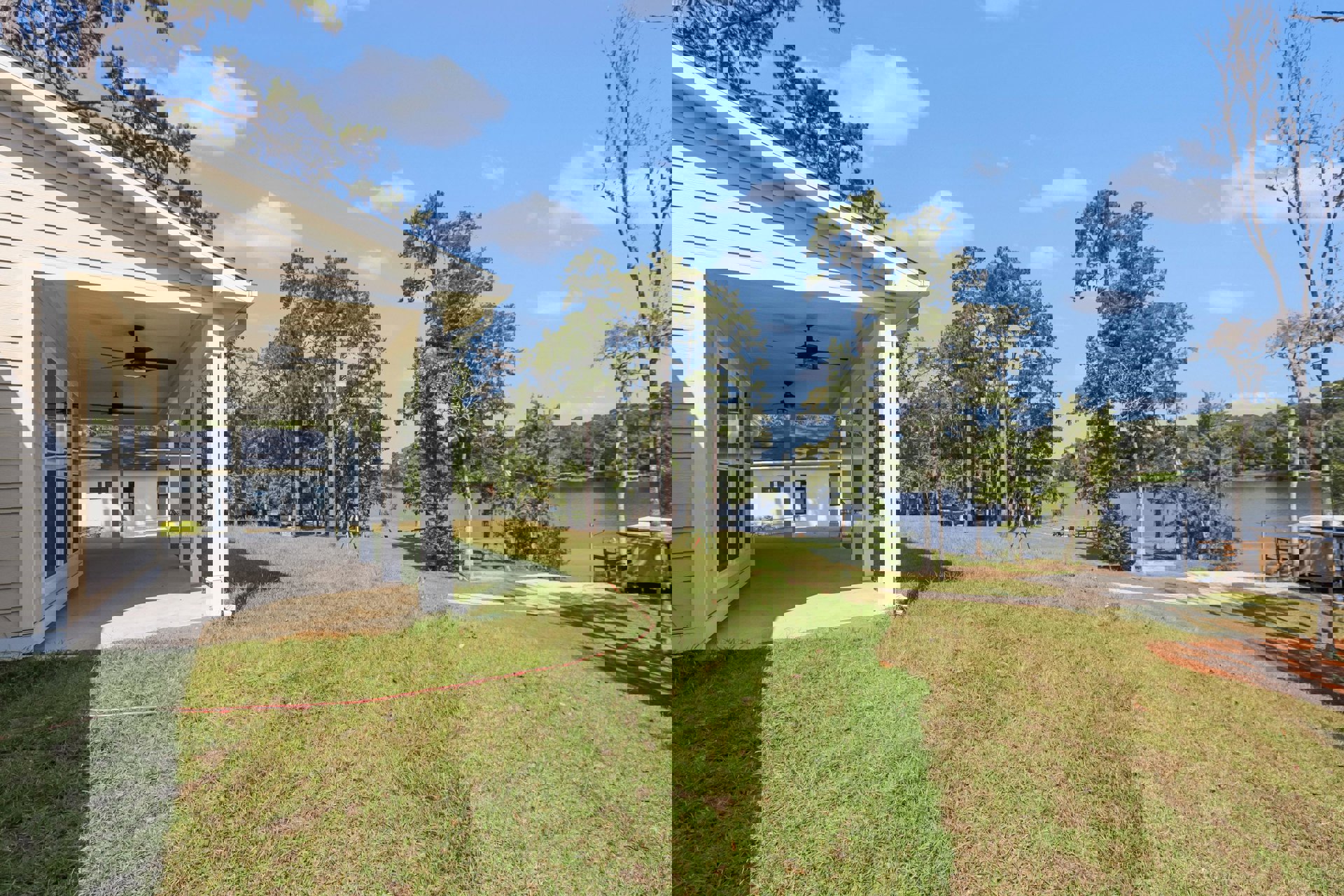 White house with covered porch, ceiling fan, and manicured lawn overlooking a lake, surrounded by leafy trees under partly cloudy sky